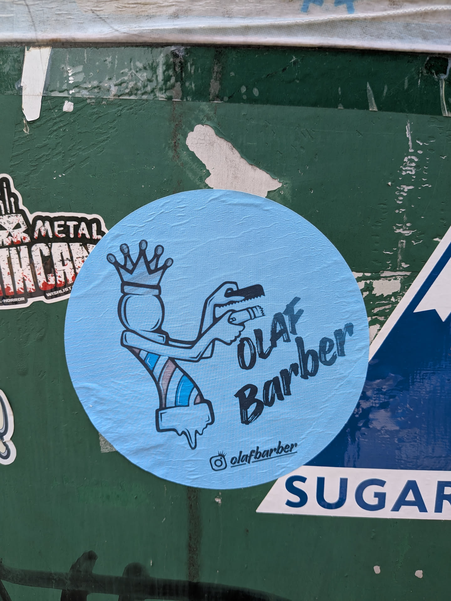 A circular light blue sticker featuring a stylized character that combines a barber pole and a crowned figure. The character has arms holding a comb and clippers. The body has diagonal stripes in blue, white, and brown, with a dripping effect at the bottom. The text 'OLAF Barber' is written in a dark blue, hand-drawn style font to the right of the character. An Instagram handle '@olafbarber' is printed at the bottom.