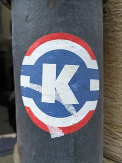 A round sticker with a white capital K in the center. The K is set against a blue background, which is circled by a white ring and then a red outer ring. The sticker is slightly damaged, with some peeling and tearing around the edges. It is affixed to a dark gray or black pole.