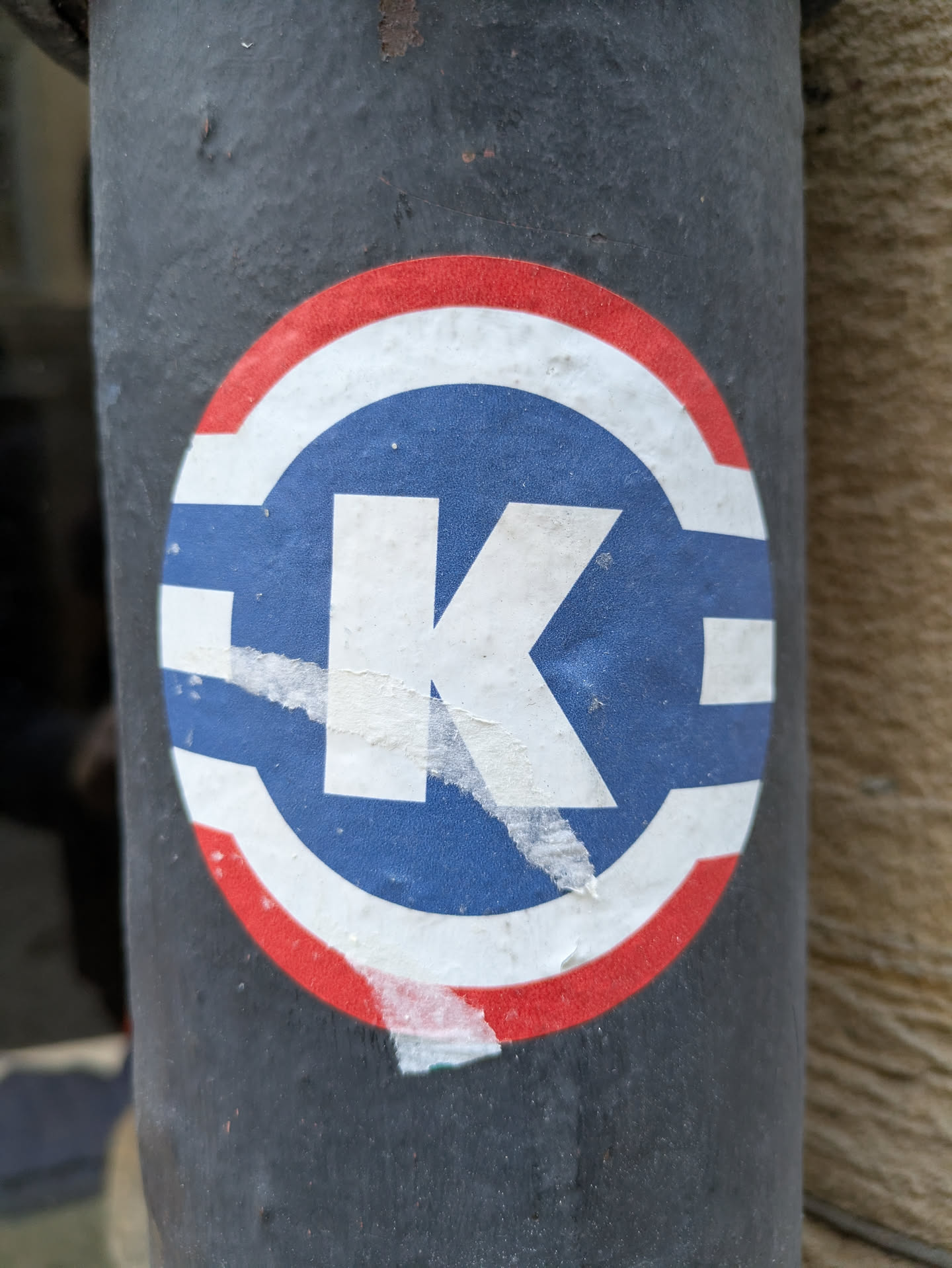 A round sticker with a white capital K in the center. The K is set against a blue background, which is circled by a white ring and then a red outer ring. The sticker is slightly damaged, with some peeling and tearing around the edges. It is affixed to a dark gray or black pole.