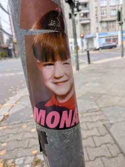 Street sticker A portrait sticker adhered to a metal lamppost depicting a retro-style photograph of a young child with auburn hair and bangs, smiling and winking one eye. The child is wearing a dark top with a prominent red collar. Spanning the lower portion of the sticker is the name 'MONA' in bold, bright pink sans-serif capital letters.