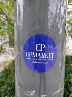 A round blue sticker with white text on a gray pole. The text on the sticker says EP UPMARKET, new & old fashion, Established 2022.