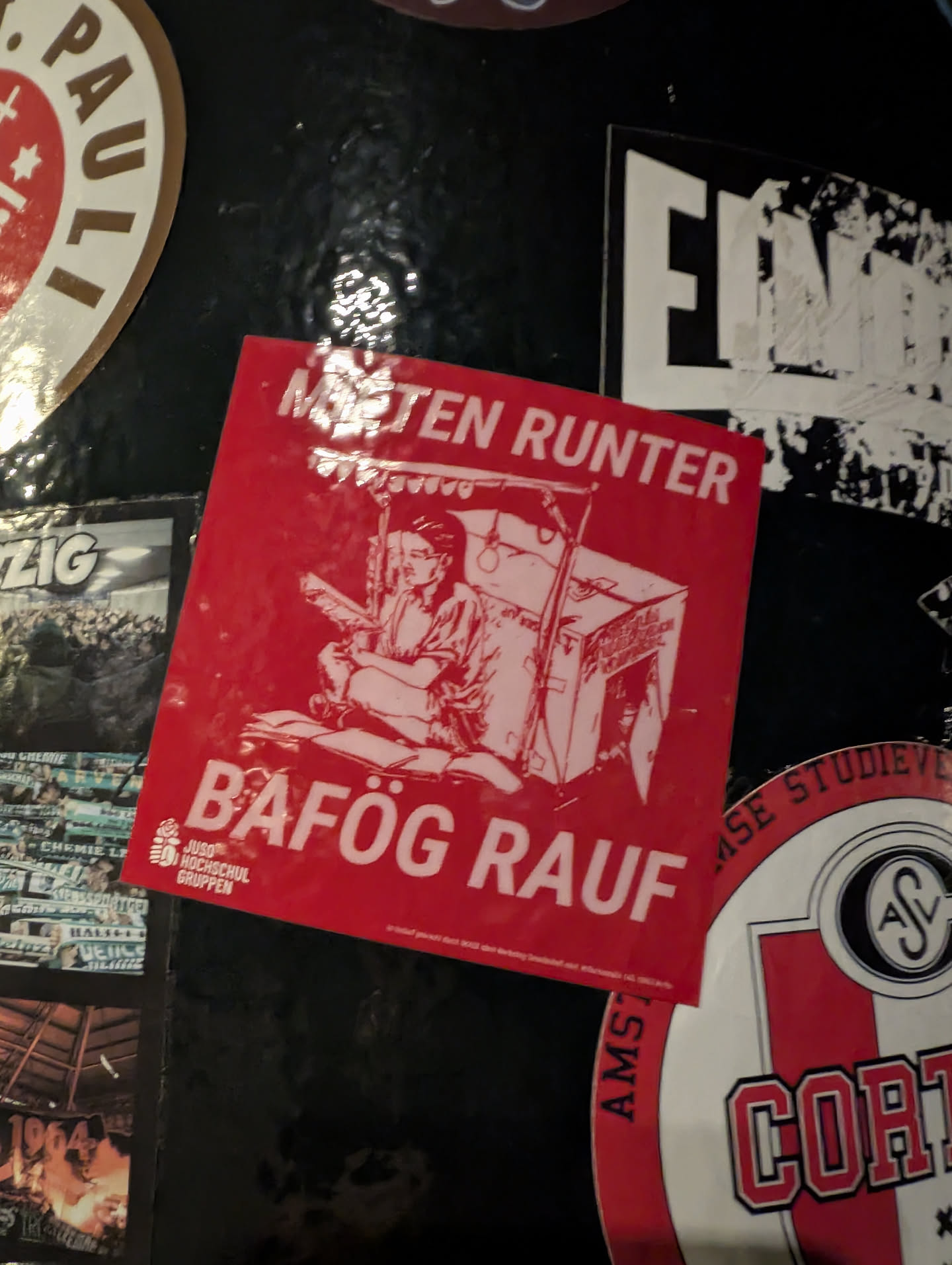 A square, vibrant red sticker featuring a white line-art illustration. The central image depicts a person, likely a student, sitting cross-legged on a stack of books, holding another book or tablet. Behind them is a makeshift structure resembling a stall or shelter, possibly constructed from cardboard boxes, with various items hanging from it, suggesting a struggle for resources. Above the illustration, in bold white uppercase letters, is the German phrase 'MIETEN RUNTER' (Rents Down). Below the illustration, also in bold white uppercase letters, is 'BÄFÖG RAUF' (BAföG Up, referring to the German student loan/grant program). In the bottom left corner, a small white logo of a rose (symbol of JUSO) is visible, accompanied by the text 'JUSO HOCHSCHUL GRUPPEN' (JUSO University Groups). The sticker has a glossy finish, reflecting light.
