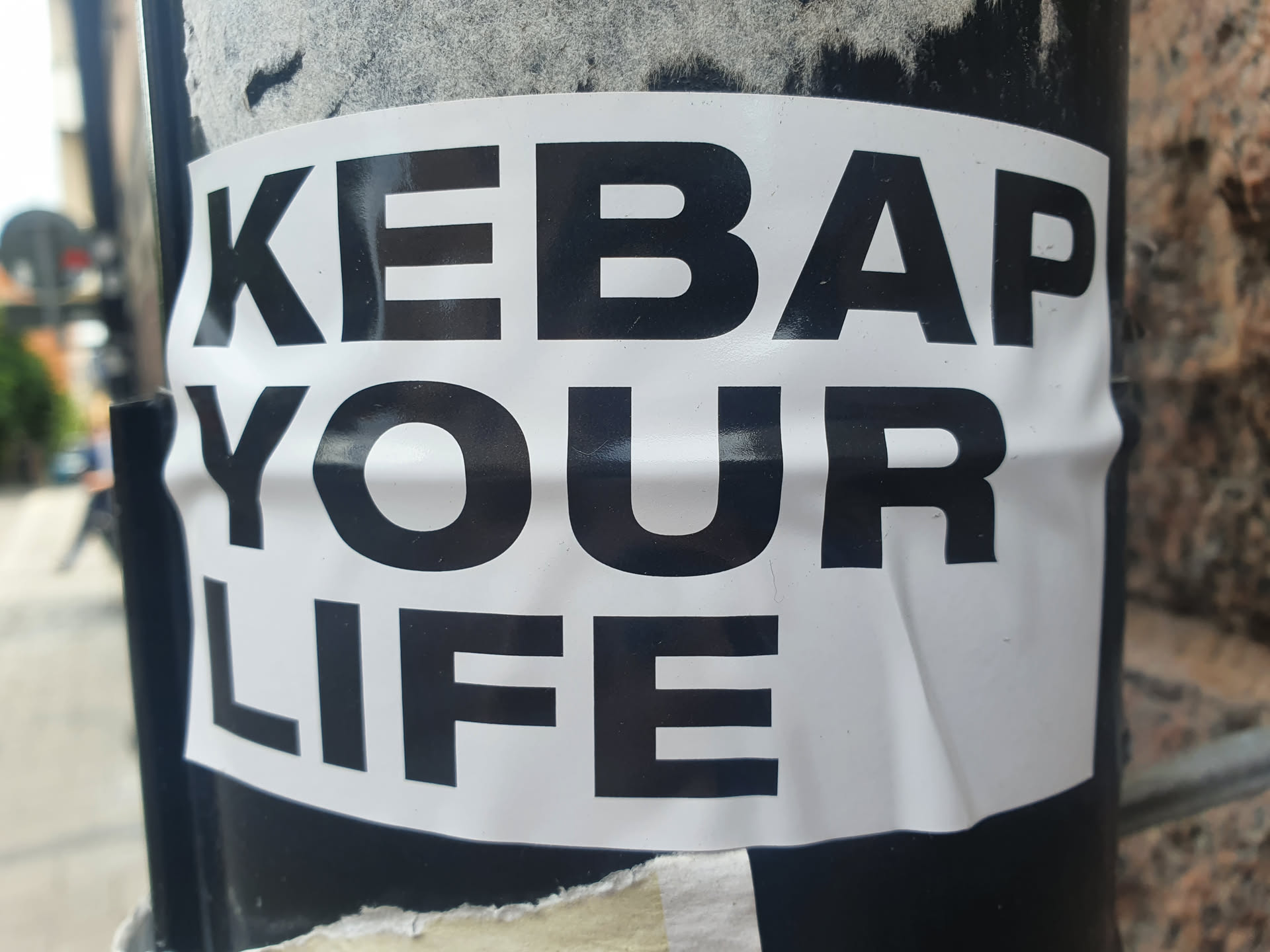 A black and white sticker with the text "KEBAB YOUR LIFE" pasted on a dark pole. The sticker is slightly wrinkled.