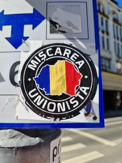 A round sticker featuring the Romanian flag and the text "Miscarea Unionista" which translates to "Unionist Movement". The sticker is attached to a pole with other stickers.