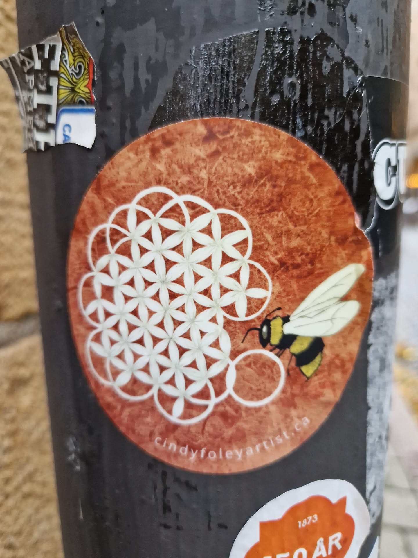 Round sticker featuring a bumblebee interacting with a Flower of Life geometric pattern on a textured reddish-brown background.  The artist's website is printed at the bottom.