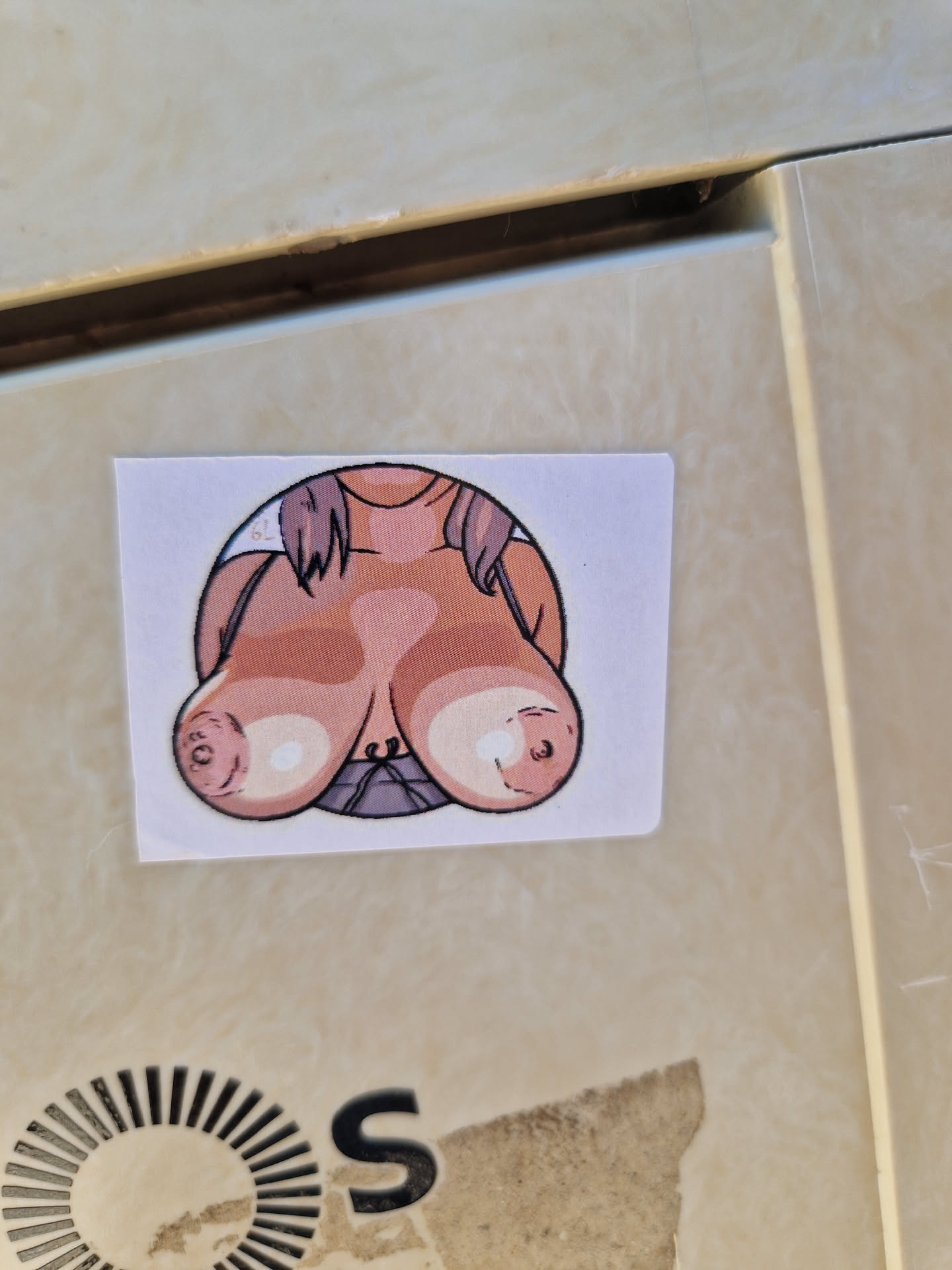 A sticker depicting a cartoon drawing of a woman's breasts. The drawing is in a circular frame on a white square sticker. The woman has light brown hair and is wearing a purple top.
