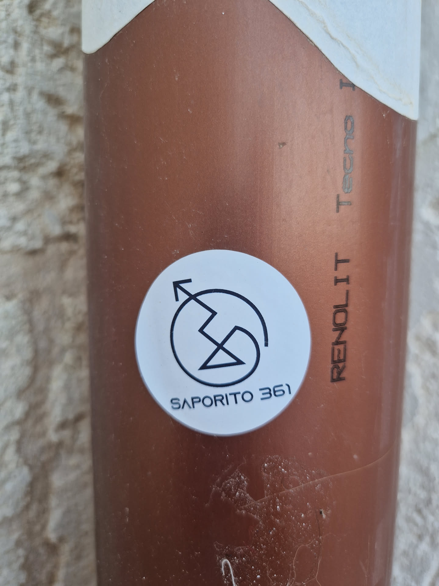 A round white sticker with a unique logo and the text 'Saporito 361'. The logo appears to be an abstract symbol possibly representing movement or energy. The sticker is affixed to a copper-colored object.