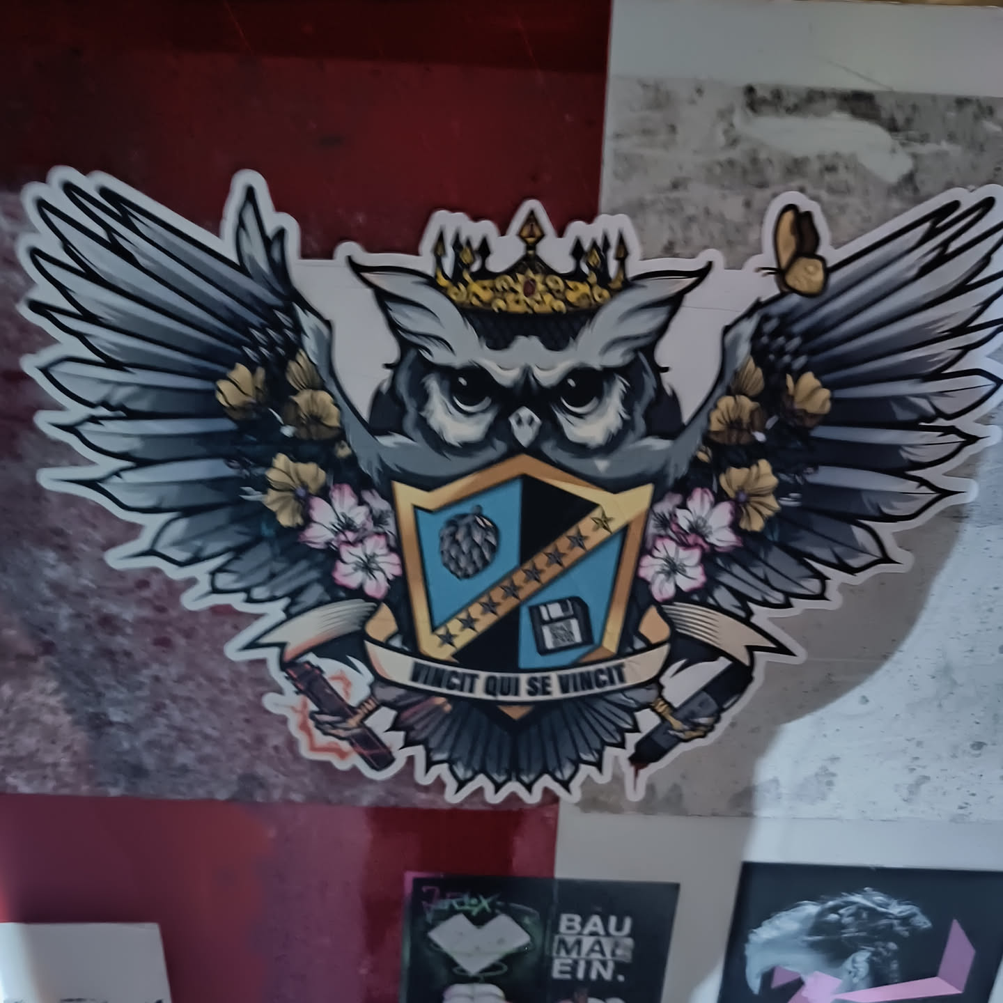 A sticker featuring an illustration of an owl wearing a crown and large wings.  The owl is positioned on a coat of arms style shield with a hop, stars, and a floppy disk.  The text below reads "Vincit qui se vincit".