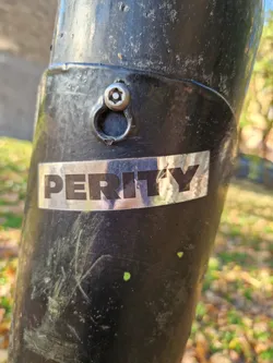 A silver Perity sticker affixed to a dark-colored metal pole outdoors. The sticker is rectangular and features the word "PERITY" in bold, uppercase lettering. The background of the sticker appears slightly reflective.