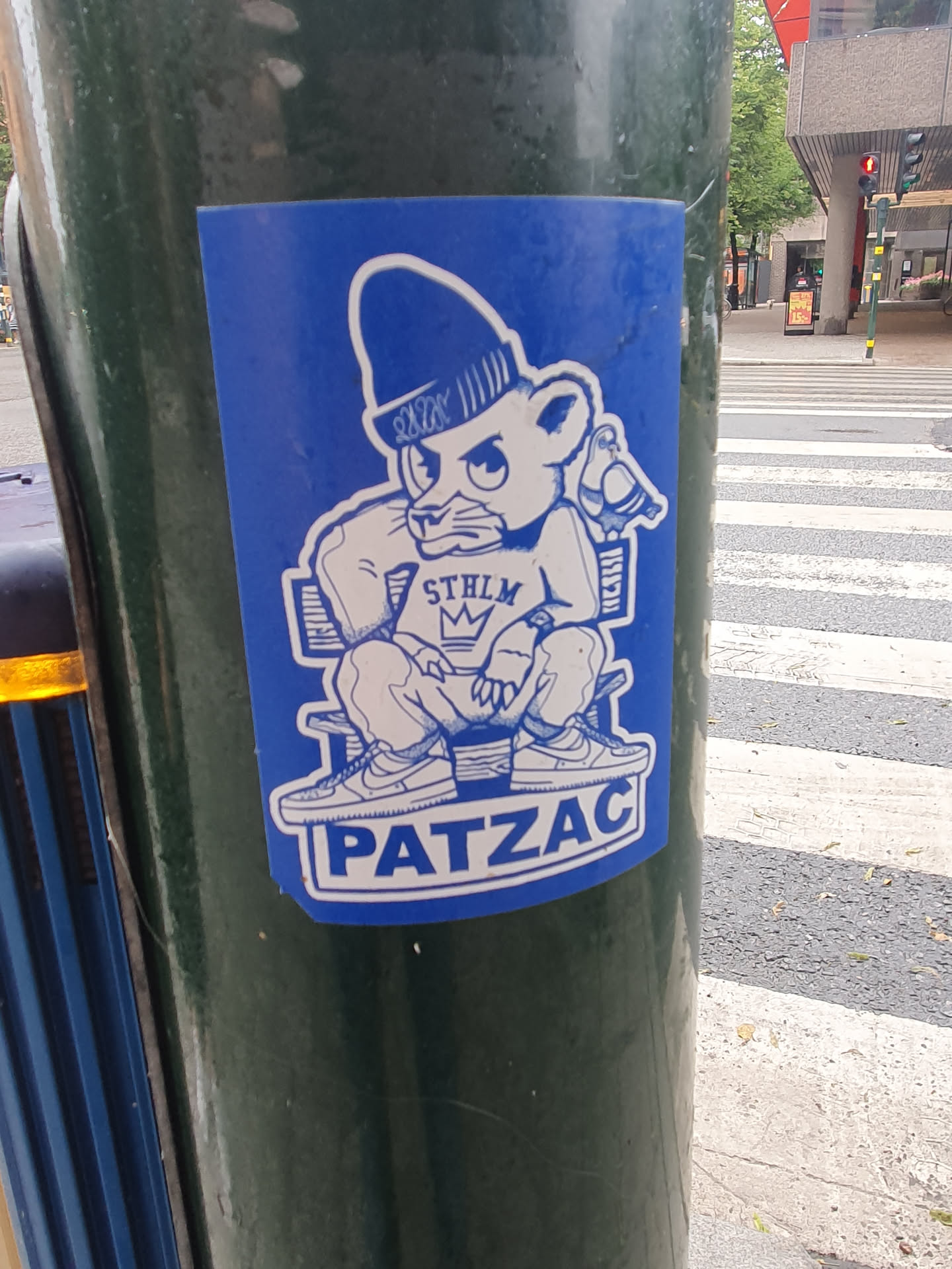 A blue sticker featuring a stylized illustration of a bear wearing a beanie and streetwear, sitting on a chair. The bear has a crown on its chest and text reading 'STHLM' (Stockholm).  The word 'PATZAC' is prominently displayed below the illustration. The art style is reminiscent of streetwear and graphic design.