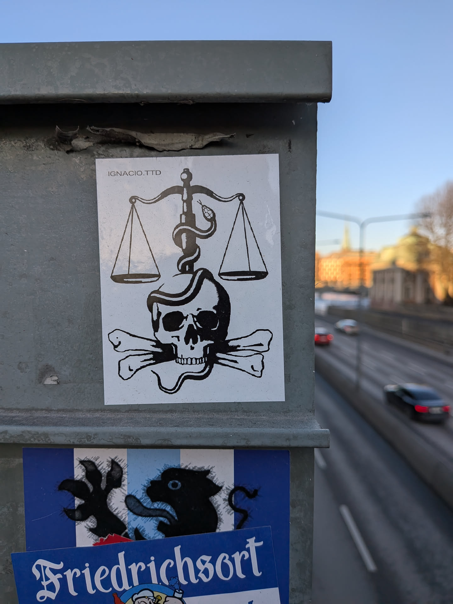 The sticker is a rectangular, black and white design. It prominently features a human skull with two crossbones positioned beneath it, in the style of a Jolly Roger. Above the skull, a classic set of scales of justice is depicted, with two empty pans. A snake is intricately wrapped around the central pillar of the scales, its head near the top pan, and its body extending down to coil around the skull. The entire illustration is rendered in bold black lines and solid black fills against a clean white background.