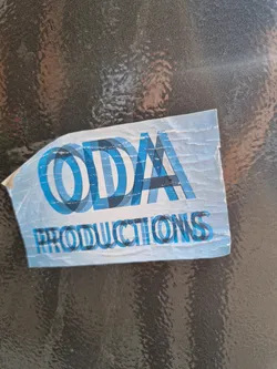 A worn-out sticker with the text 'ODA Productions' in a stylized, overlapping font. The sticker is light blue with dark blue text and is stuck on a dark gray surface. The sticker appears to be made of vinyl.