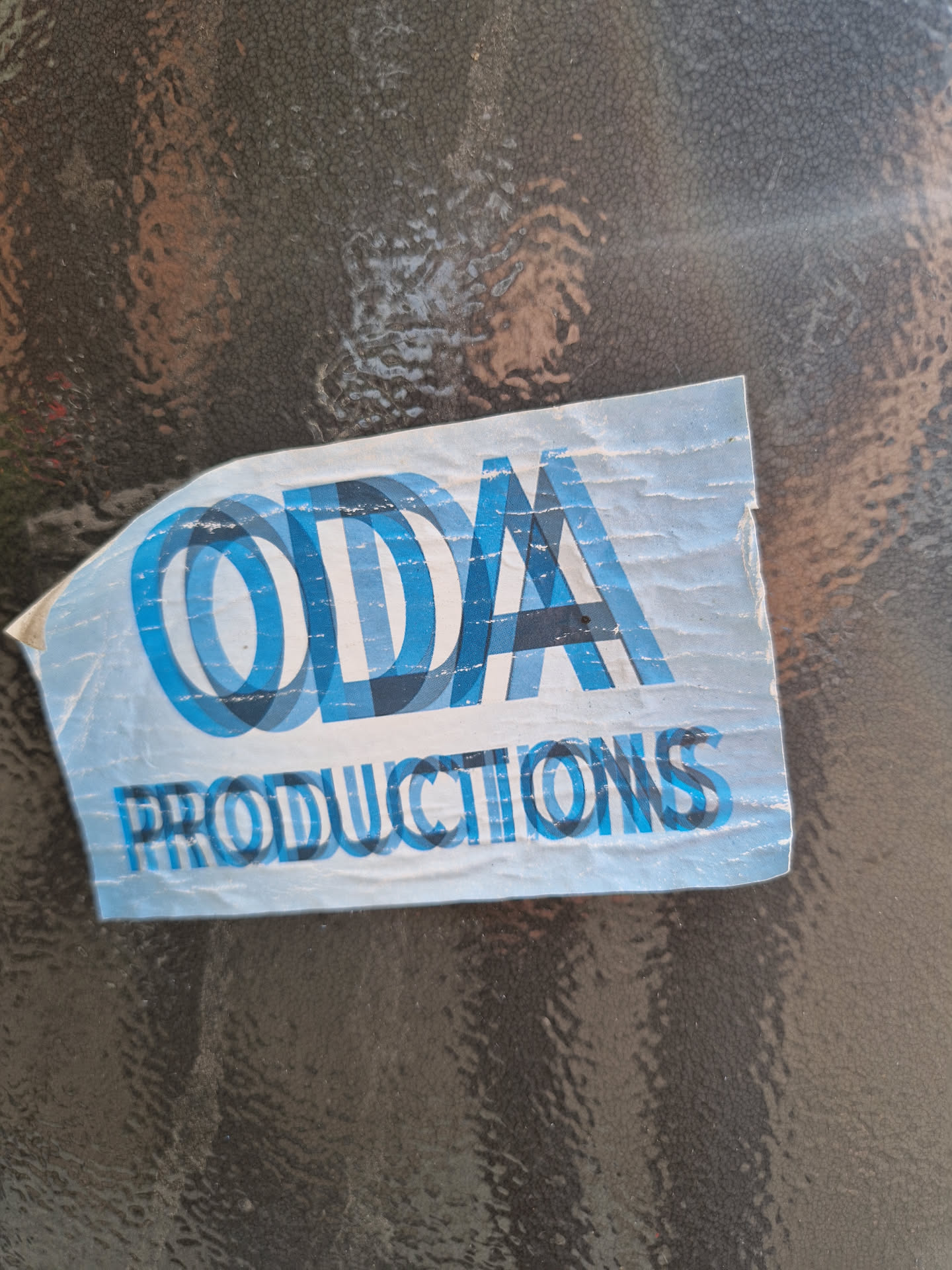 A worn-out sticker with the text 'ODA Productions' in a stylized, overlapping font. The sticker is light blue with dark blue text and is stuck on a dark gray surface. The sticker appears to be made of vinyl.