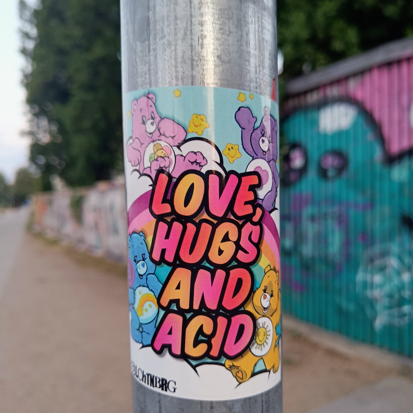 Care Bears themed sticker with a rainbow and the text "Love, Hugs and Acid". The sticker is on a metal pole in an urban setting.