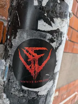 A circular sticker featuring a highly stylized, intricate, and somewhat jagged symbol in a vibrant blood-red color against a solid black background. The symbol appears to be composed of three main, claw-like or wing-like elements extending outwards from a central point, with a textured, almost corroded or dripping effect. Below the central symbol, in smaller red capital letters, is a website address. The sticker is affixed to a weathered, dark pole, surrounded by remnants of other torn paper and grime.