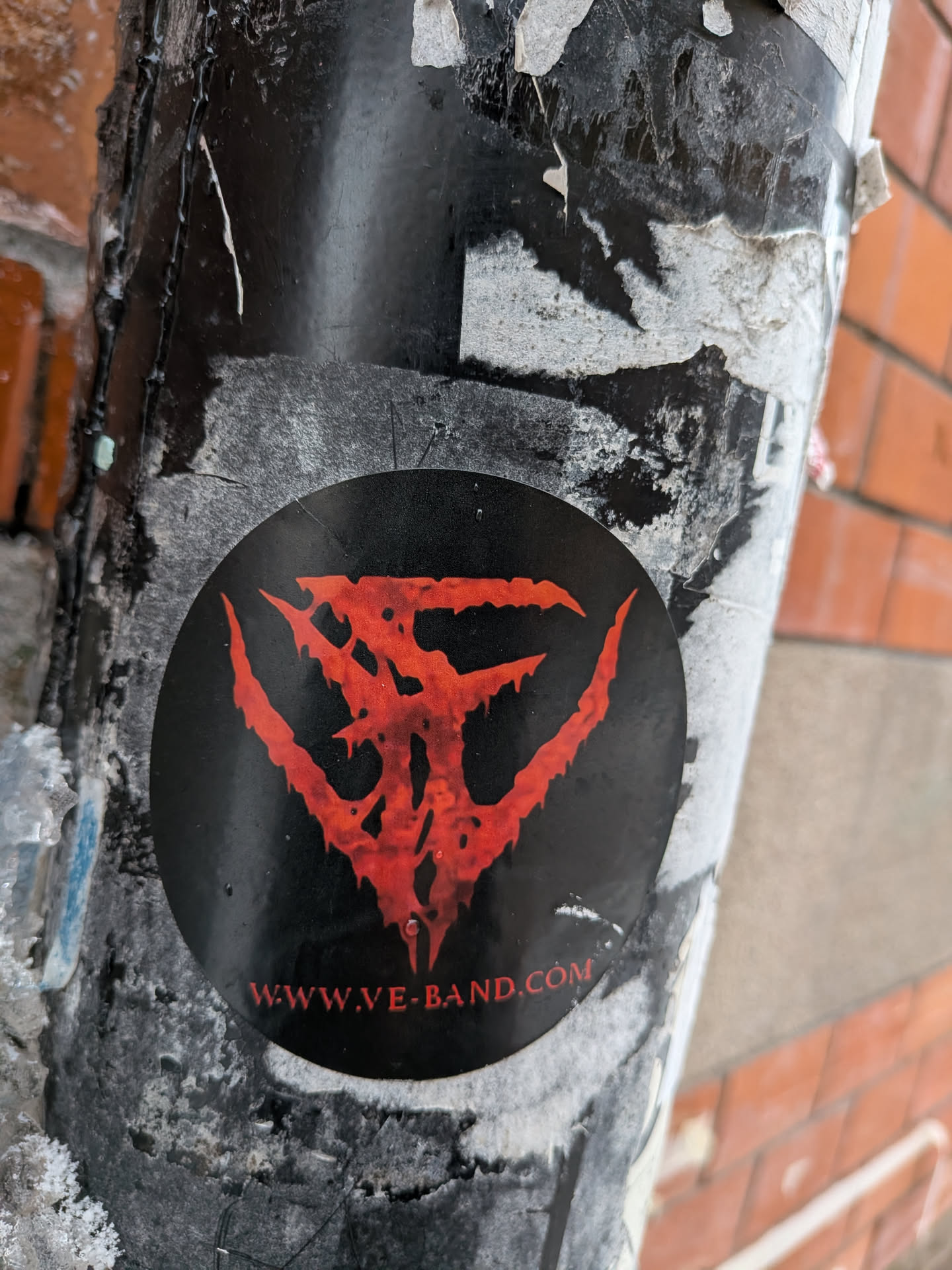 A circular sticker featuring a highly stylized, intricate, and somewhat jagged symbol in a vibrant blood-red color against a solid black background. The symbol appears to be composed of three main, claw-like or wing-like elements extending outwards from a central point, with a textured, almost corroded or dripping effect. Below the central symbol, in smaller red capital letters, is a website address. The sticker is affixed to a weathered, dark pole, surrounded by remnants of other torn paper and grime.