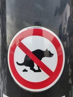 Round sticker with a red circle and a diagonal red line over a black silhouette of a dog pooping.  The text at the top says "www.stickers-enzo.nl".