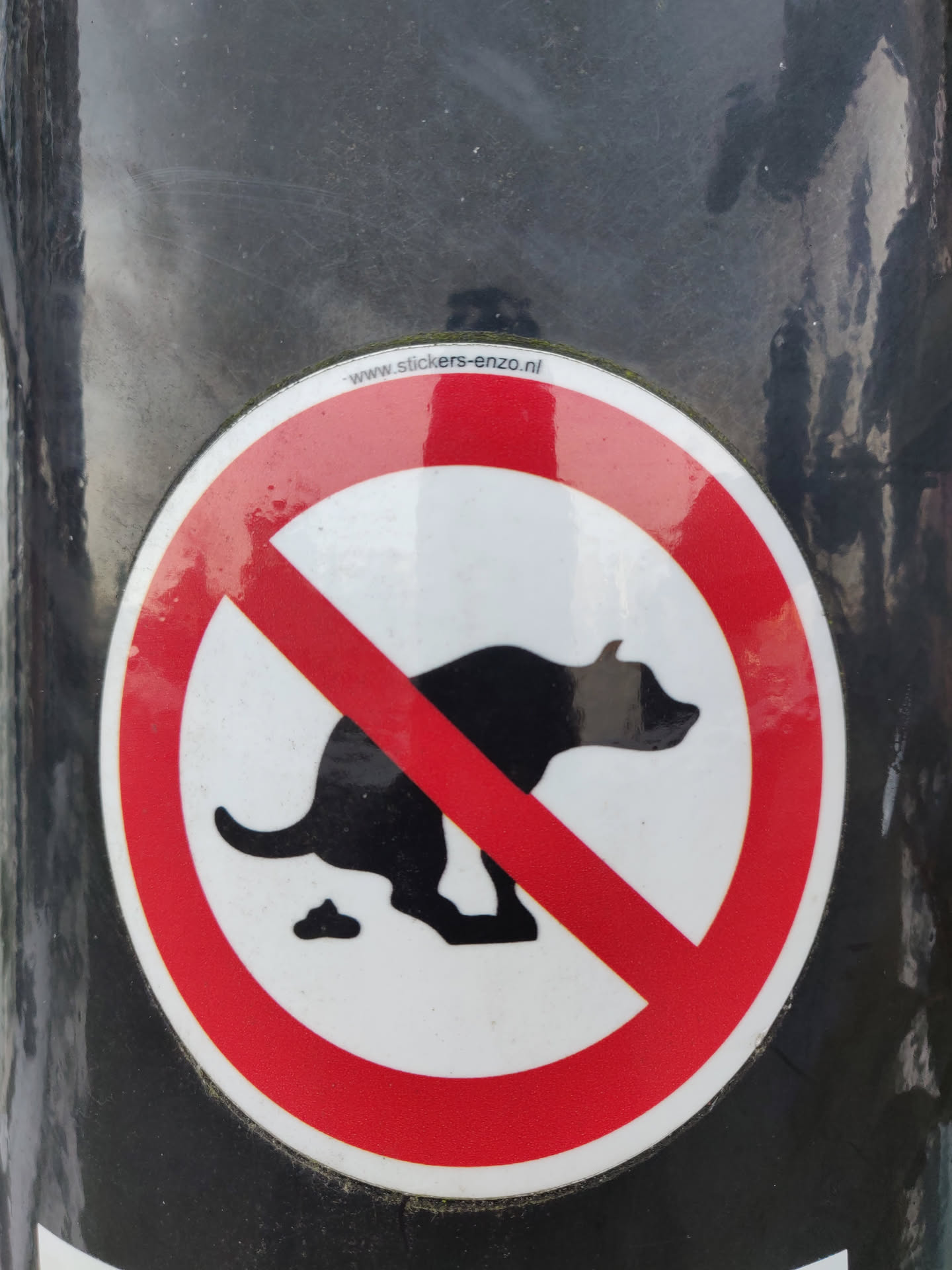 Round sticker with a red circle and a diagonal red line over a black silhouette of a dog pooping.  The text at the top says "www.stickers-enzo.nl".