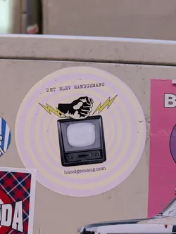 Street sticker The sticker is a circular design featuring a vintage television set with a fist punching down on it, accompanied by lightning bolts. The background consists of concentric circles in a pale lavender and white color scheme. The text "DET BLEV HANDGEMANG" is arched above the image, and "handgemang.com" is printed below.