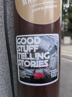 This sticker features the text "GOOD STUFF TELLING STORIES" in bold, white letters above an image of train tracks stretching into the distance within a tunnel. Below the image, there's a red signature and a QR code. The sticker promotes storytelling, investigating, and archiving cultural knowledge and values.