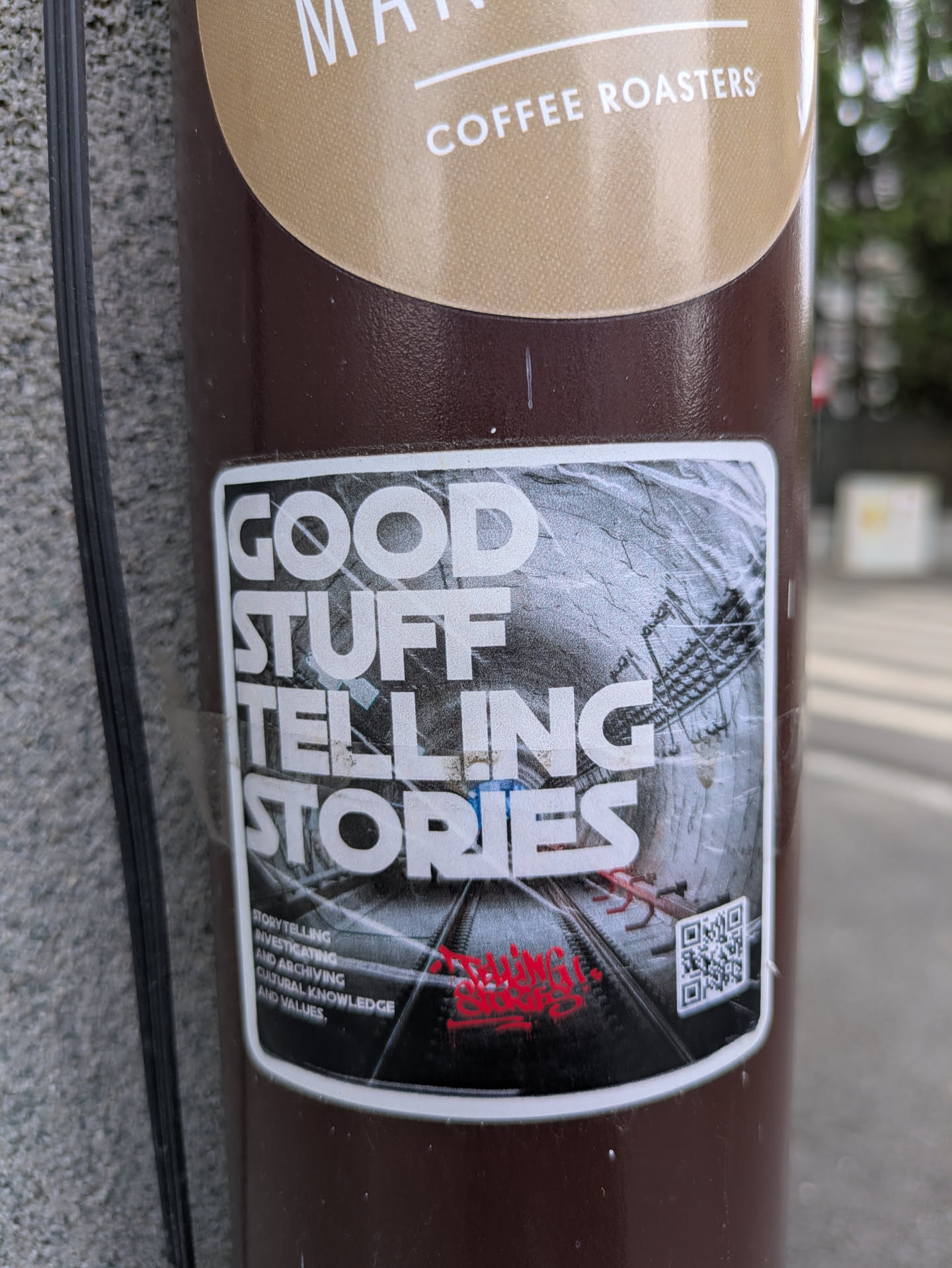 This sticker features the text "GOOD STUFF TELLING STORIES" in bold, white letters above an image of train tracks stretching into the distance within a tunnel. Below the image, there's a red signature and a QR code. The sticker promotes storytelling, investigating, and archiving cultural knowledge and values.