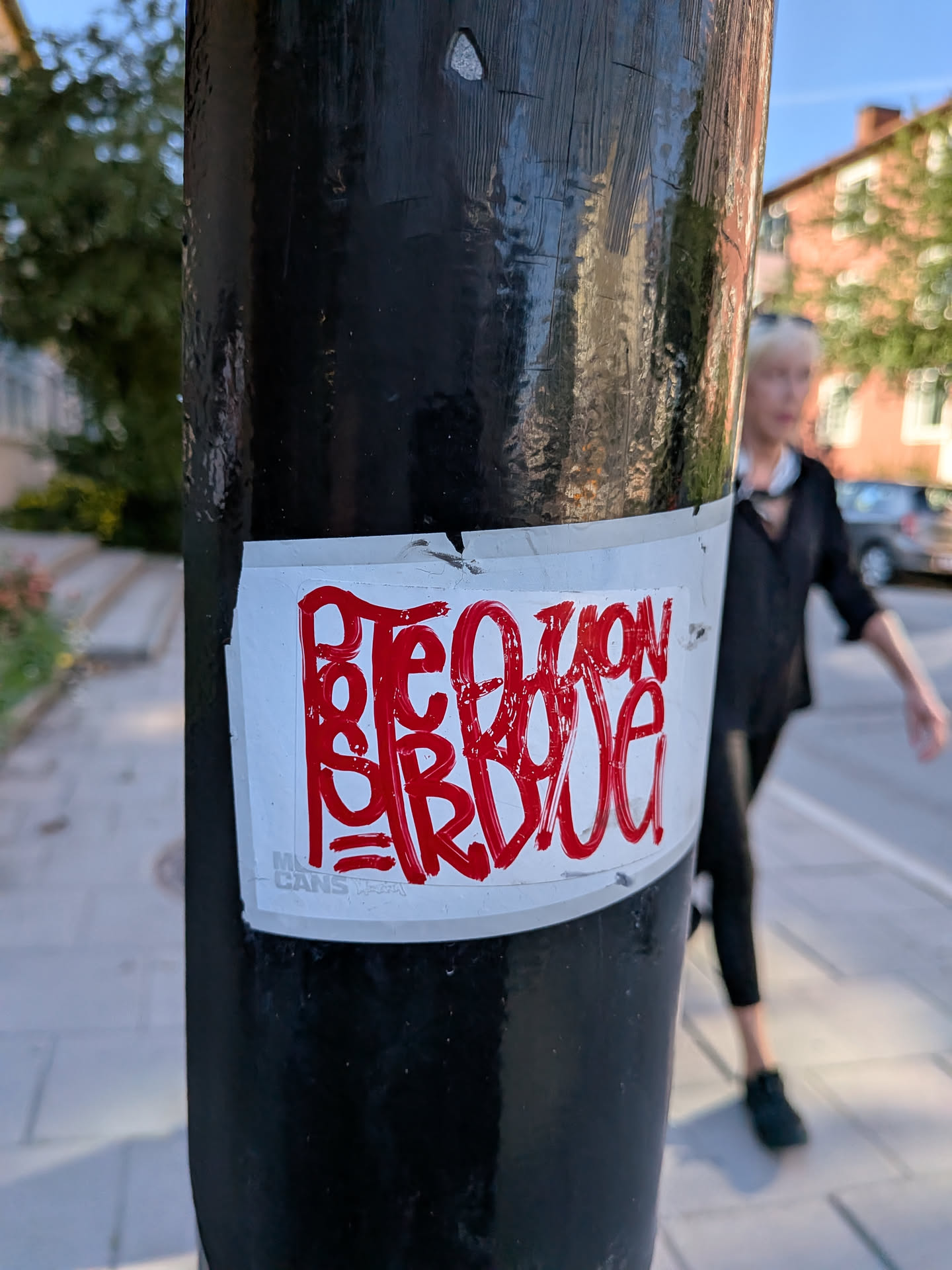 The sticker is rectangular with a white background and is attached to a dark, cylindrical object, likely a pole. The lettering is done in a bold, flowing red marker with a slight sheen, giving it a graffiti-like appearance. The text reads 'Peter Bon', with the letters connected and stylized. There appears to be a small logo or mark in the bottom left corner of the sticker.
