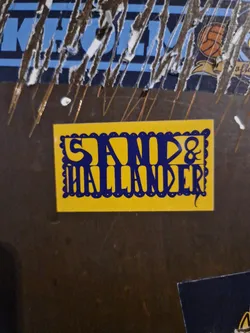 Close-up of a yellow sticker with the text "Sand & Hallander" in dark blue. The sticker has a decorative border.