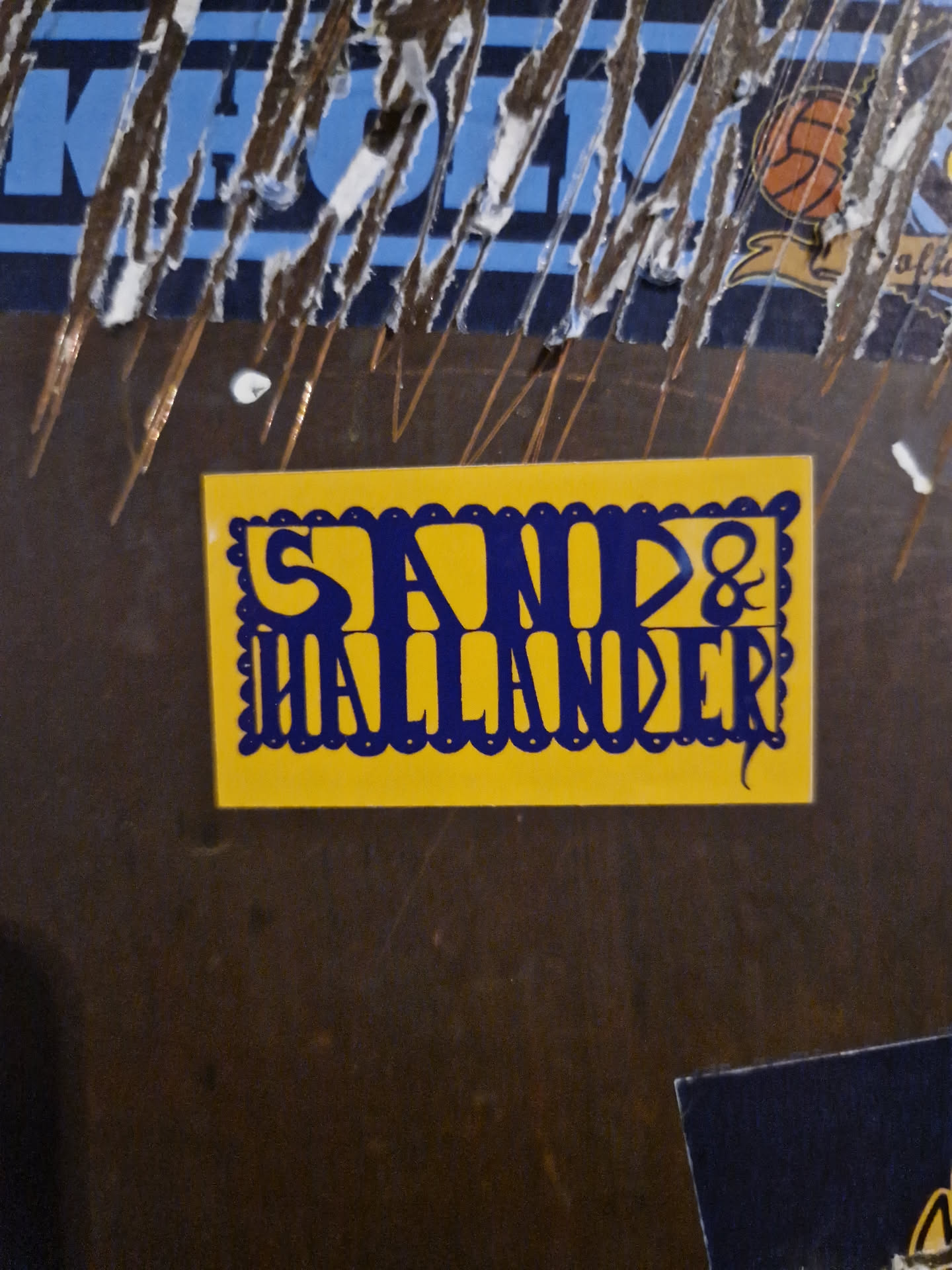 Close-up of a yellow sticker with the text "Sand & Hallander" in dark blue. The sticker has a decorative border.