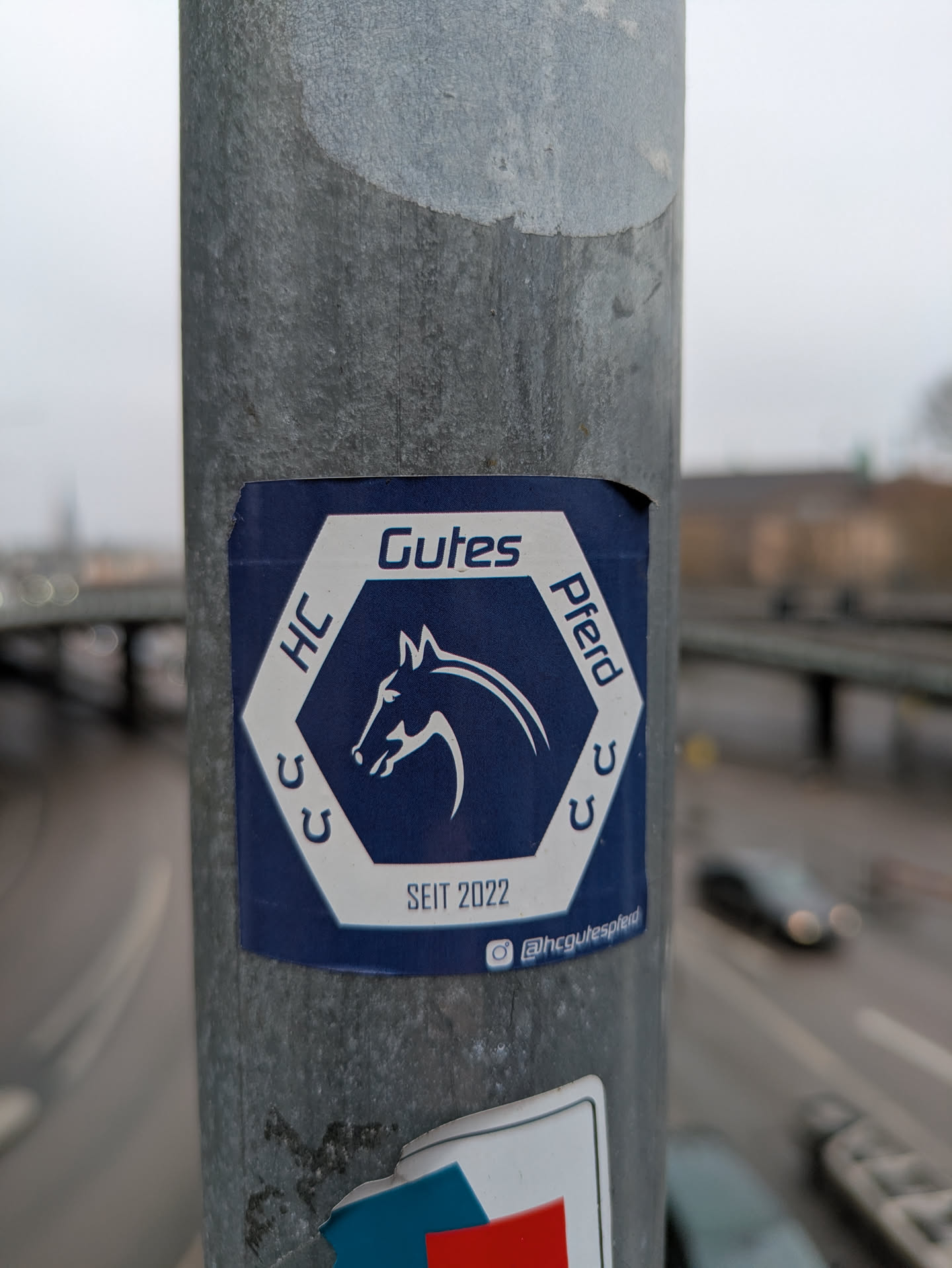A sticker on a light pole advertises 'HC Gutes Pferd' (HC Good Horse) since 2022. The sticker is a dark blue hexagon with white text and a horse head silhouette inside. It also features horseshoes and an Instagram handle.