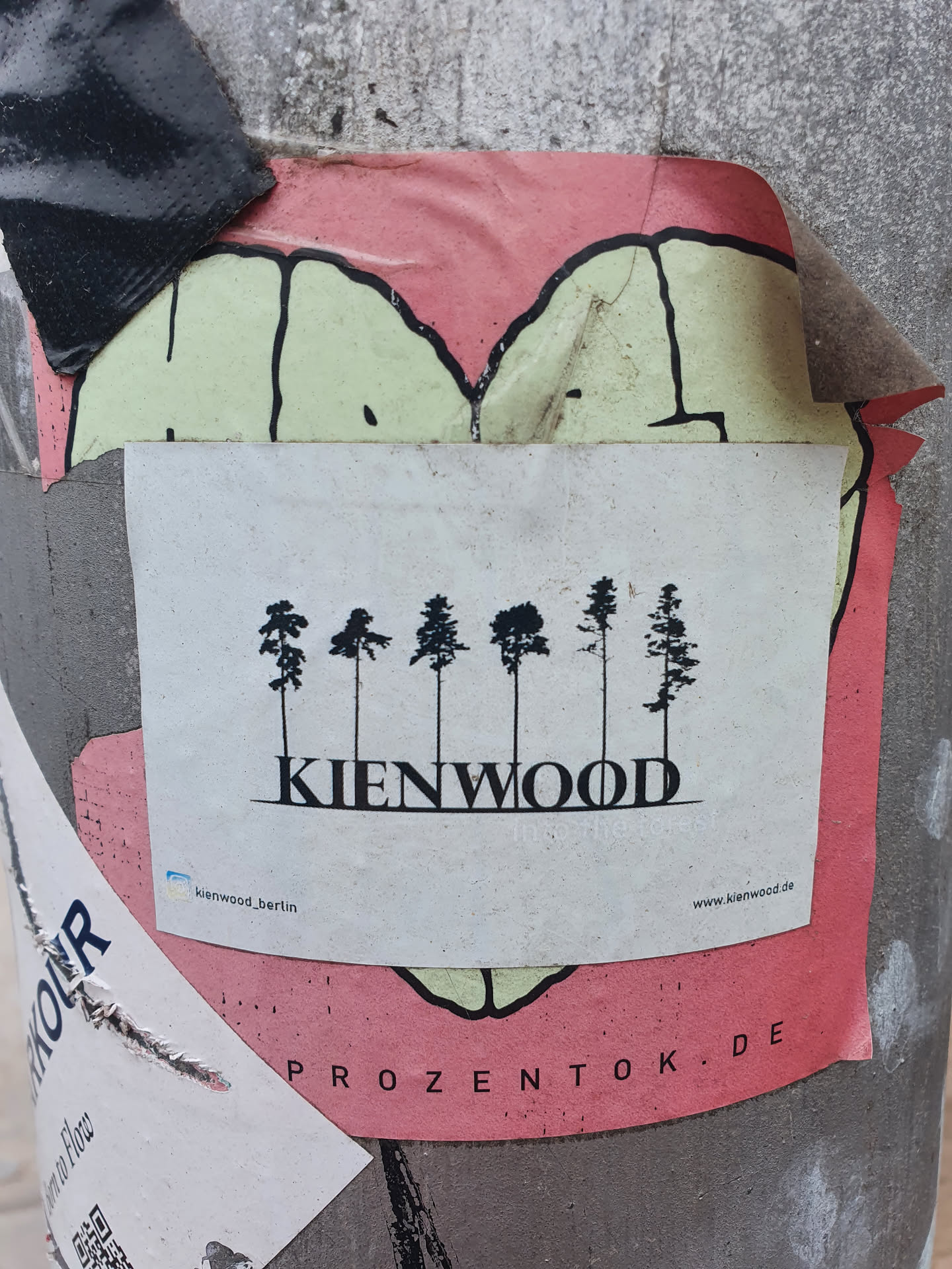 A minimalist sticker featuring a row of five stylized pine trees and the word 'KIENWOOD' in a sans-serif font.  The sticker is affixed to a pole with other stickers and graffiti visible in the background. A website address is visible at the bottom of the sticker.