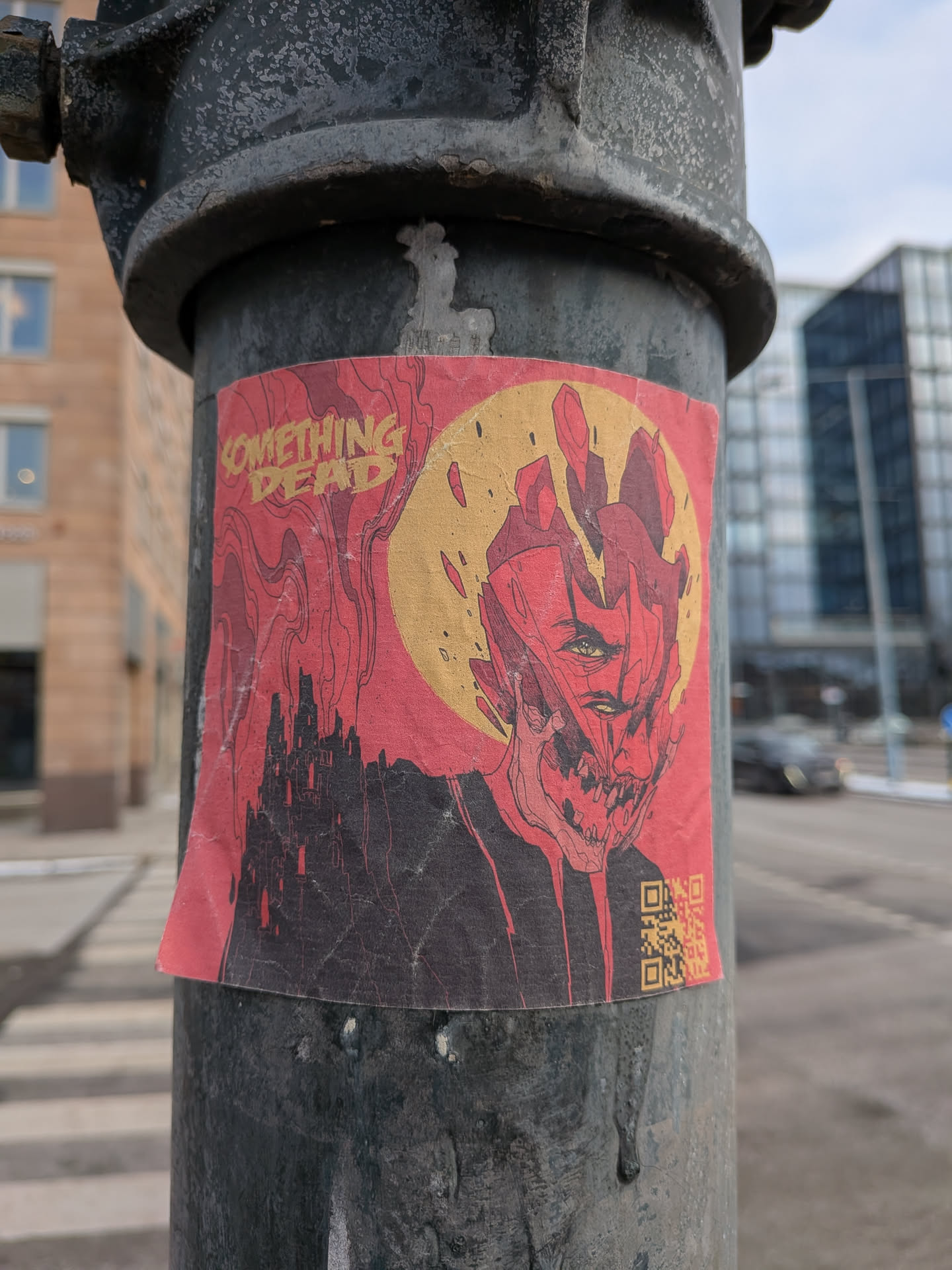 A rectangular sticker adhered to a dark metal pole, showing signs of wear with creases and slightly peeling edges. The artwork is predominantly red and black with yellow/gold accents. On the left side, a jagged, dark castle silhouette rises against a red background with swirling, smoke-like lines. The right side features a highly stylized, grotesque, and fragmented face, possibly demonic or monstrous, rendered in red and black outlines with some red fill. Its eyes are a piercing yellow/greenish hue. Behind the face is a large, rough-edged yellow/gold circle, resembling a moon or sun. In the top left corner, the text "SOMETHING DEAD" is written in a distressed, yellow/gold font. A small QR code, also in yellow/gold on a black background, is visible in the bottom right corner.