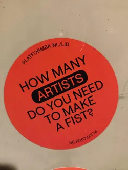 Round red sticker with a question written in black: How many artists do you need to make a fist?  There is also a website address on the sticker.