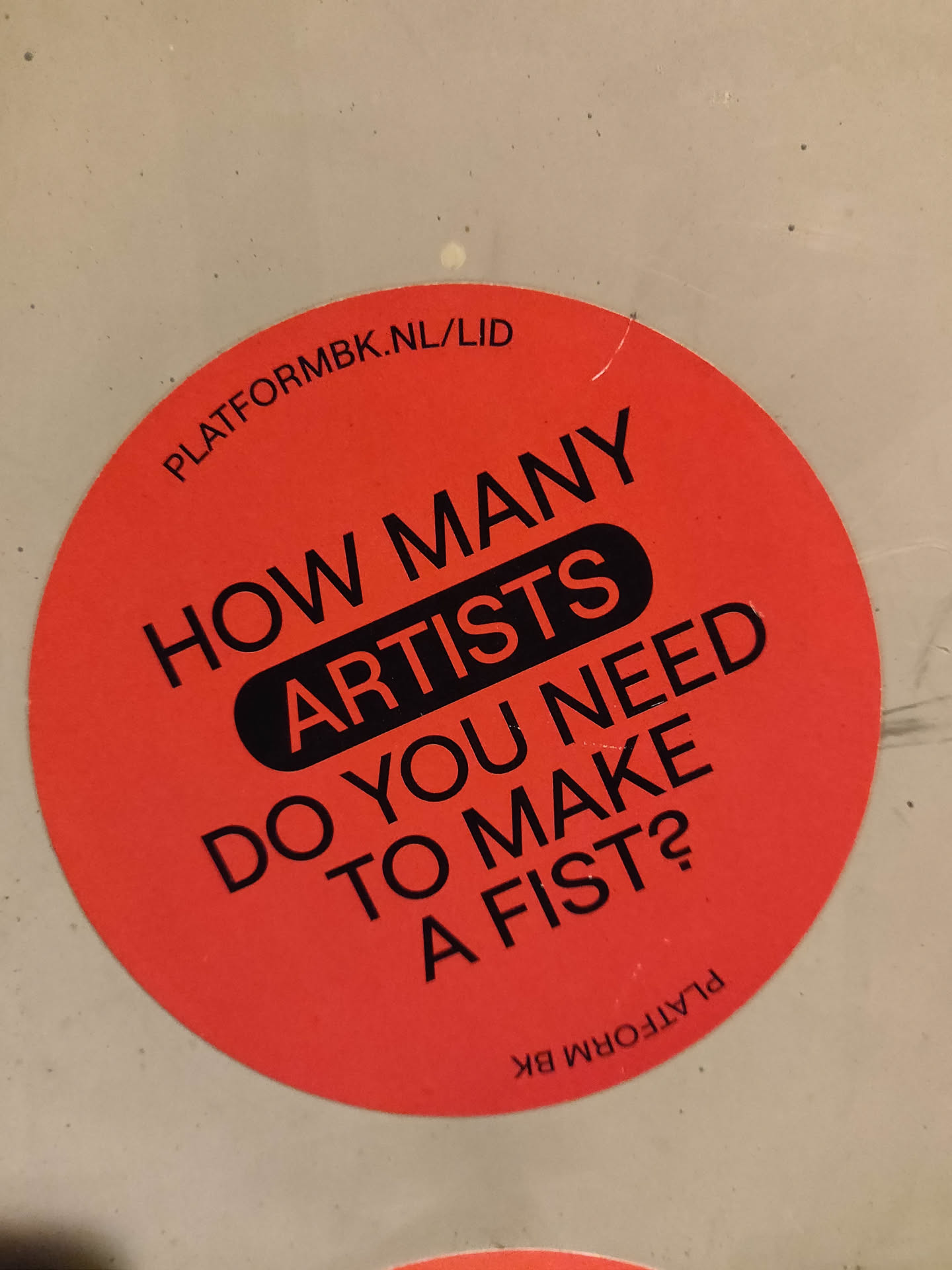 Round red sticker with a question written in black: How many artists do you need to make a fist?  There is also a website address on the sticker.