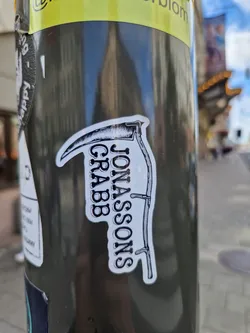 Black and white sticker with an illustration of a scythe and the text Jonassons Grabb. The sticker is placed on a dark gray pole.