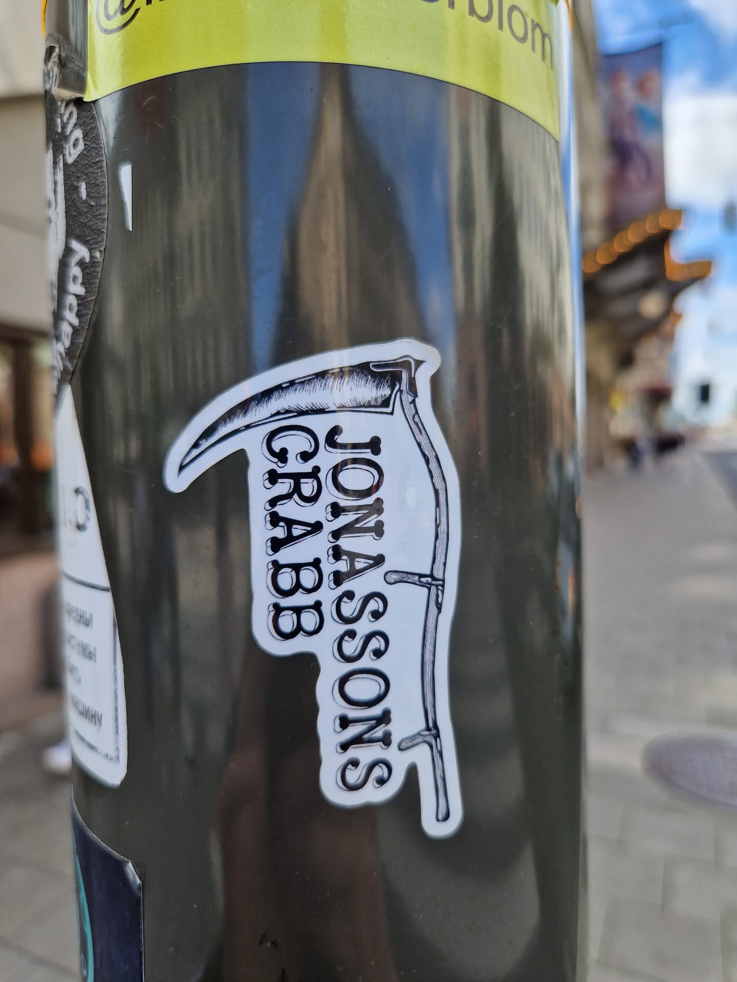 Black and white sticker with an illustration of a scythe and the text Jonassons Grabb. The sticker is placed on a dark gray pole.
