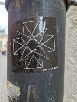 A black sticker with a geometric design. The design features intersecting lines forming triangles and other shapes. The text "TRPTYCH.COM" is visible at the bottom of the sticker.