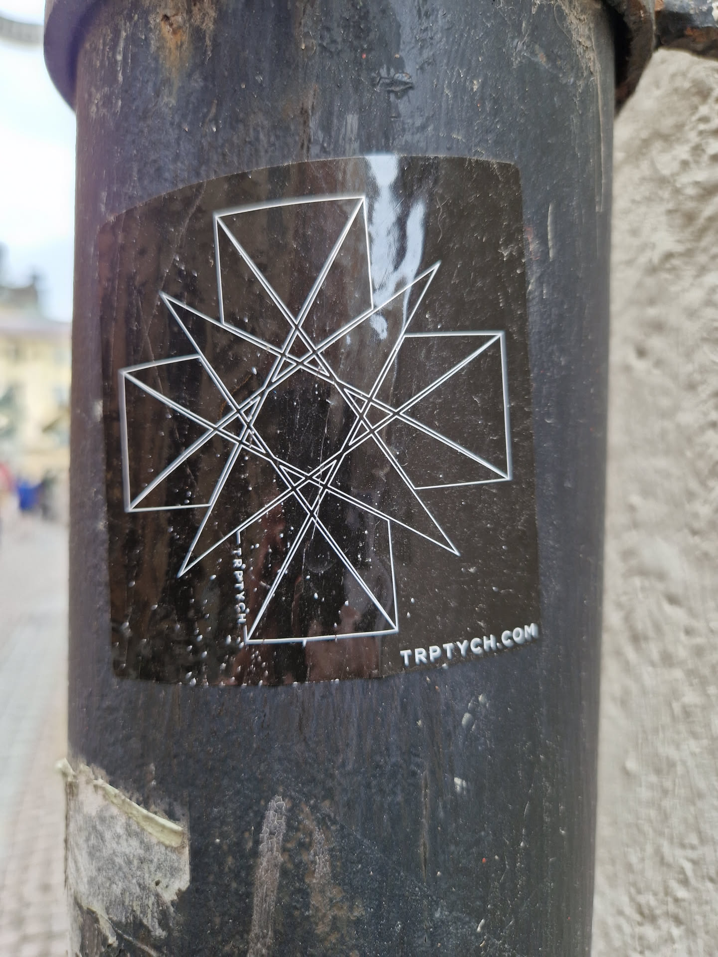 A black sticker with a geometric design. The design features intersecting lines forming triangles and other shapes. The text "TRPTYCH.COM" is visible at the bottom of the sticker.