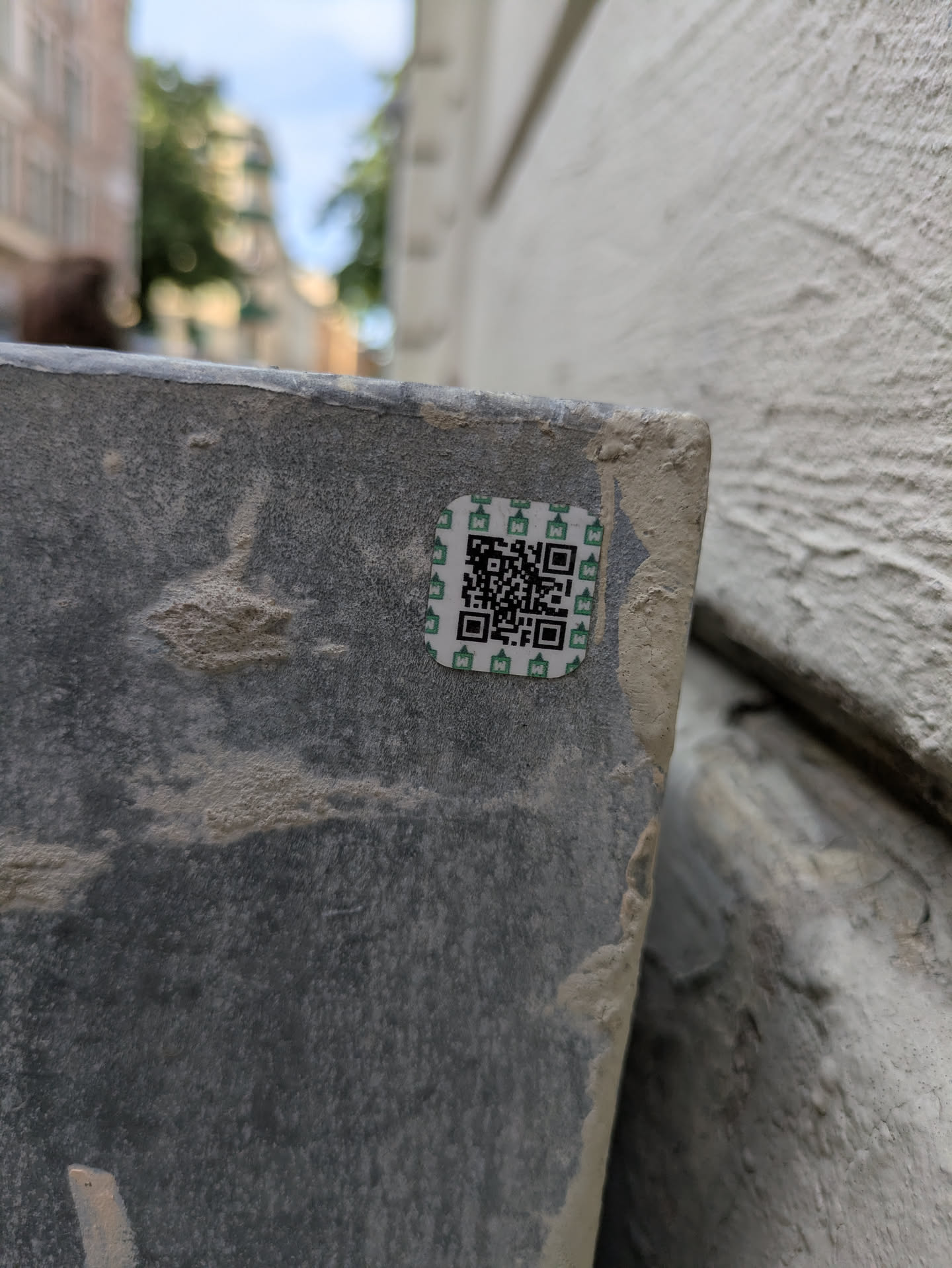 The sticker is square-shaped with a white background and a green border. It features a black QR code in the center, surrounded by a repeating pattern of small, stylized green icons that resemble buildings or pagodas. The sticker appears to be adhered to a rough, textured surface, possibly concrete or stone, with some dirt and wear visible.