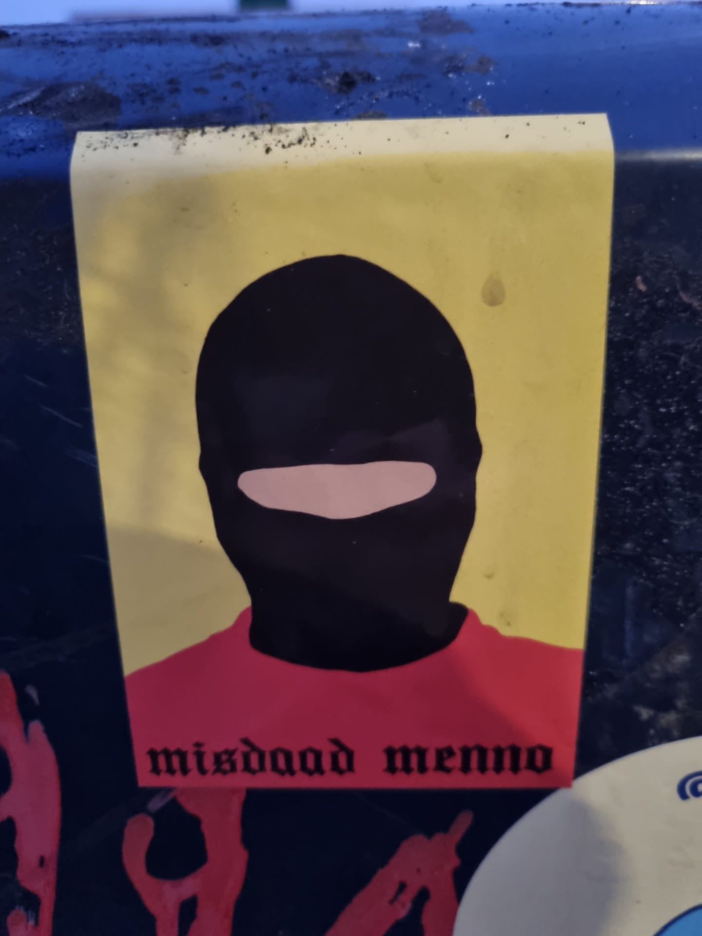 A sticker depicting a person wearing a balaclava with the text 'misdaad menno' at the bottom. The figure is shown from the chest up and is wearing a red shirt. The background is yellow.