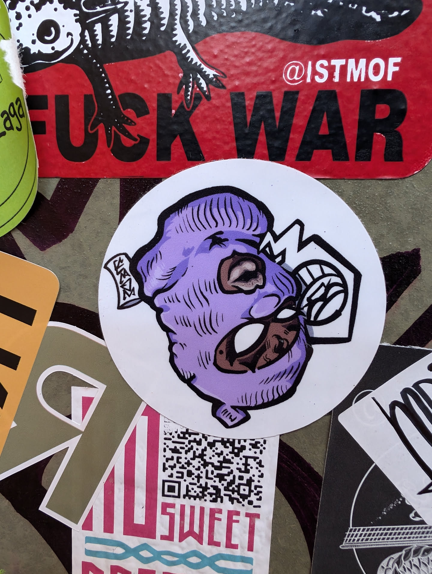 Round sticker featuring an illustration of a character wearing a purple balaclava. The style is cartoonish and hand-drawn, with visible brushstrokes and shading. The character's face is partially visible, with dark skin tones and intense eyes.  The background of the sticker is white.