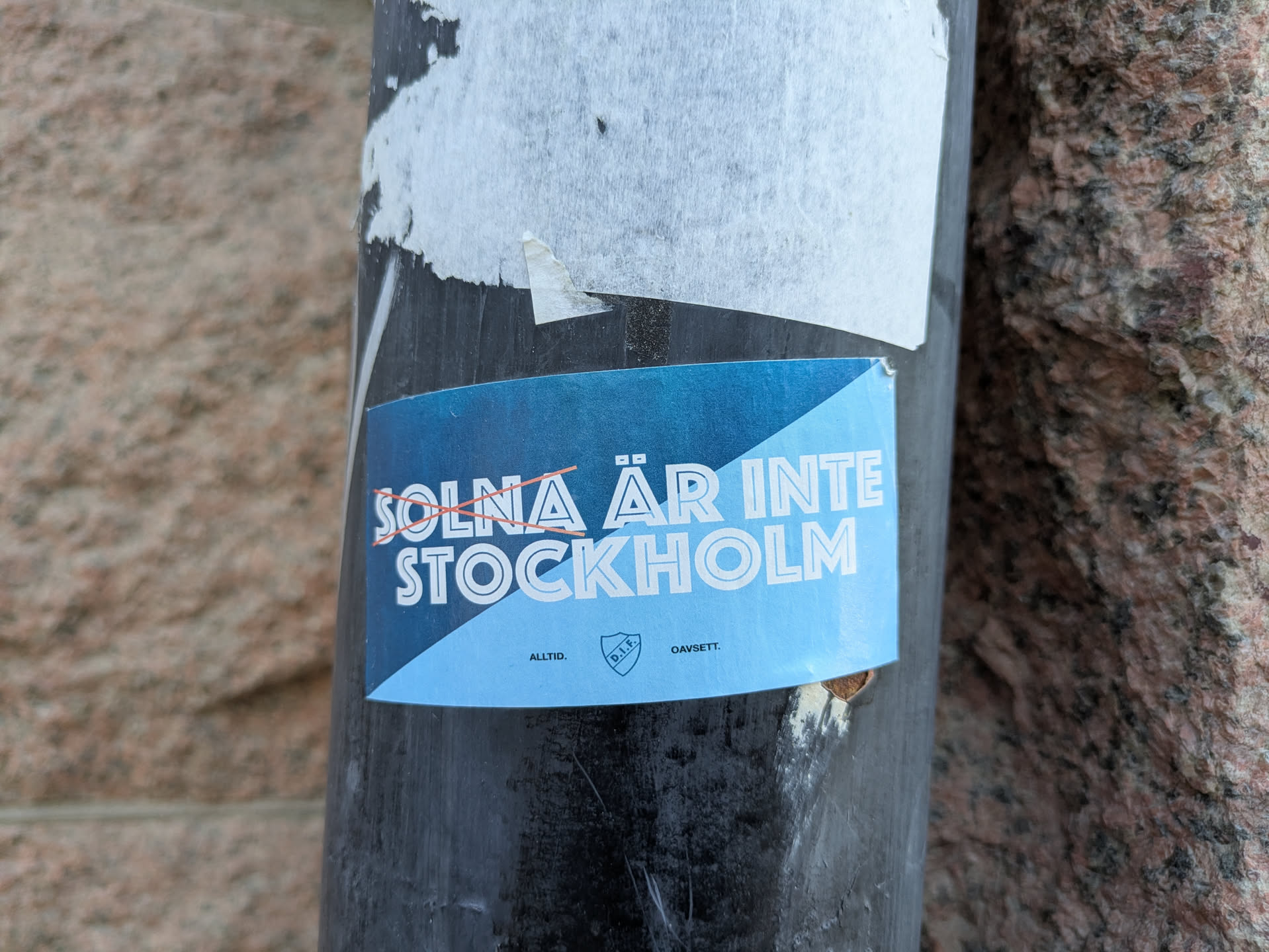 The sticker is primarily light blue with white text and a shield emblem. The text "SOLNA ÄR INTE STOCKHOLM" is prominently displayed across the top portion, with an orange line crossed through "SOLNA". Below this, the word "STOCKHOLM" is written in larger letters. At the bottom, there are smaller text elements "ALLTID." and "GAVSETT." flanking a shield with "D.I.F." inside. The sticker has a slightly curved, possibly oval or rounded rectangular shape, and is affixed to a dark, cylindrical surface. The edges of the sticker show some wear and tear, indicating it has been applied for some time.