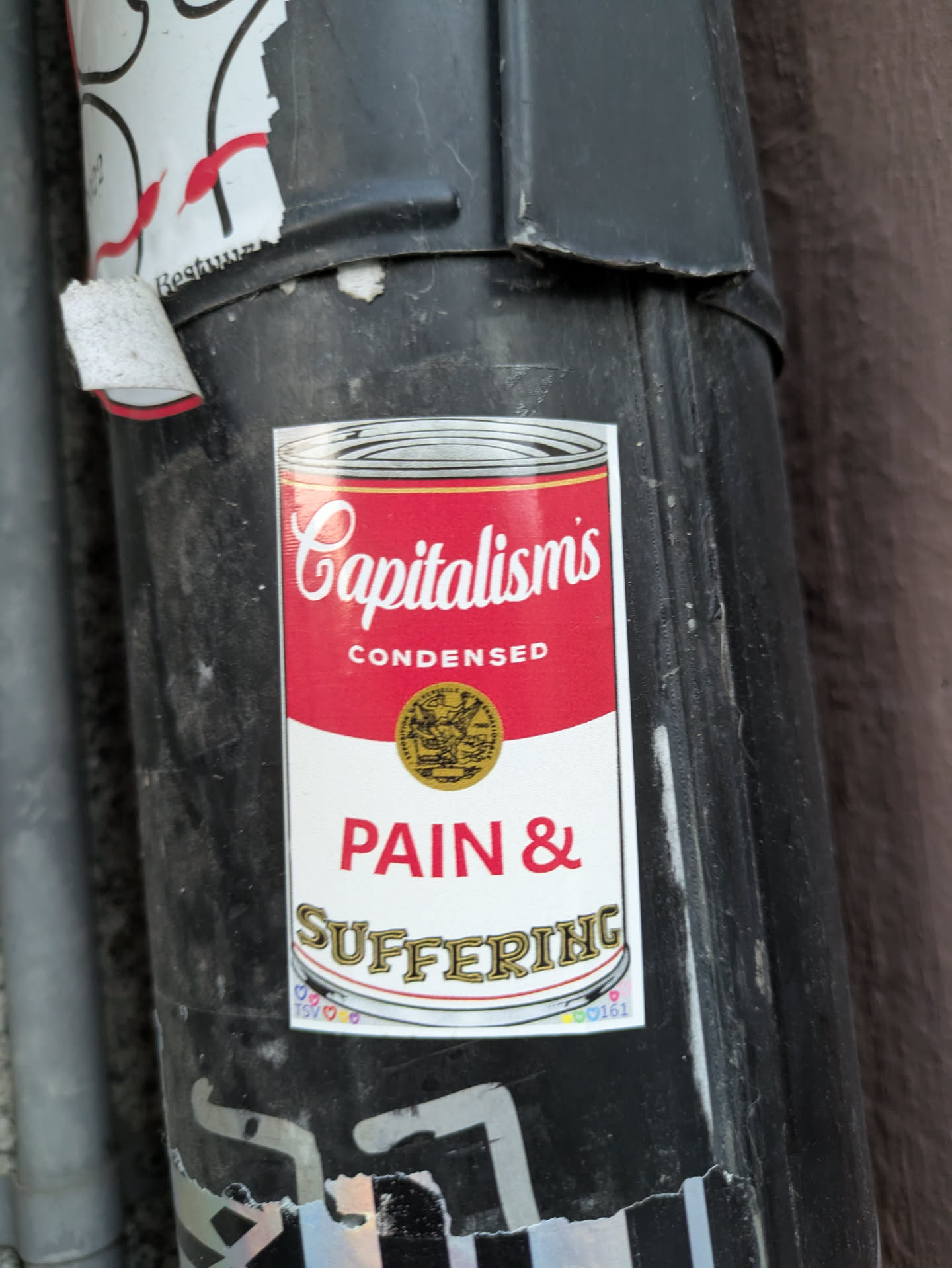 The sticker is designed to mimic the iconic Campbell's Soup can label, but with a critical message about capitalism. The top section features the word "Capitalism's" in a stylized, cursive font, followed by "CONDENSED" in a block font. Below this, a gold circular emblem with an embossed image is present. The main body of the sticker displays "PAIN & SUFFERING" in large, bold, red capital letters. The bottom of the sticker has a wavy banner with the word "SUFFERING" in a decorative, gold-colored font. There are also small, colorful heart icons and some numbers printed in the lower right corner of the sticker.