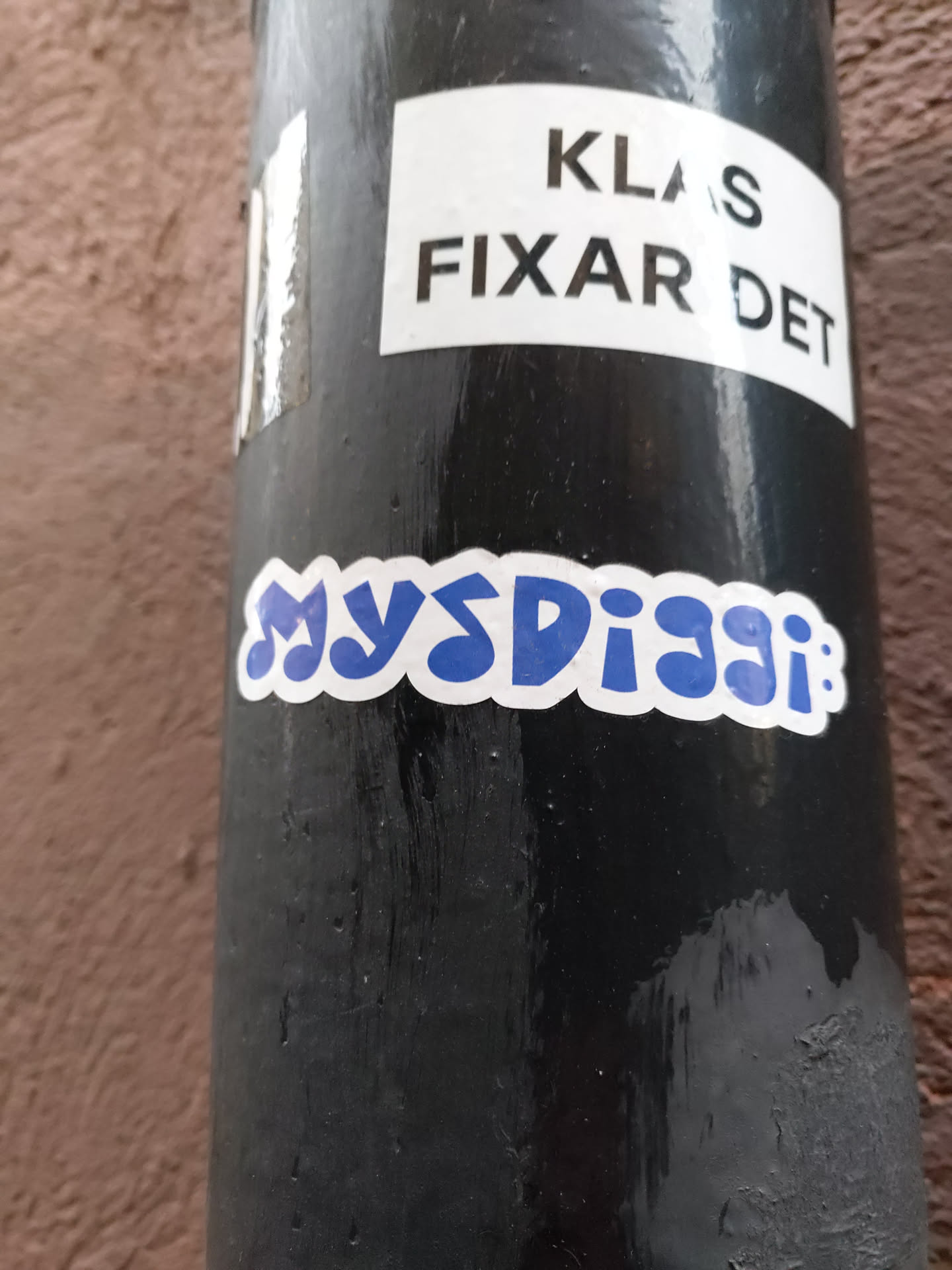 A blue sticker with the word MysDisgi in a stylized font. It's stuck on a black pole, and another sticker that says 'Klas Fixar Det' is visible above it.