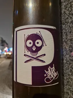 A sticker featuring a stylized skull with pencils forming a crossbones design. The skull has large, round eyes and is encircled by radiating lines, giving it a whimsical feel. The color scheme is primarily dark purple and white.  A graffiti-style signature is visible on the bottom right corner.