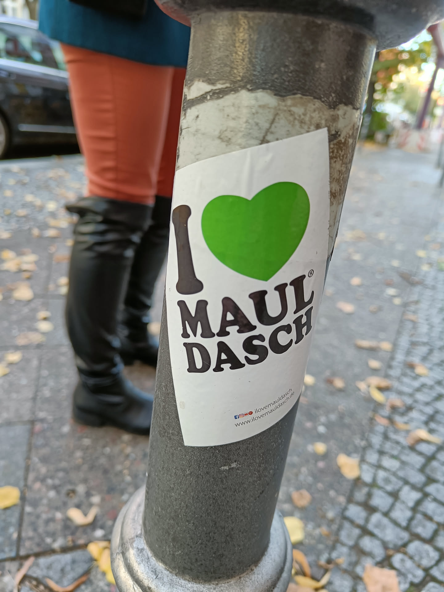 A sticker with a green heart and the text "I ♥ MAUL DASCH" is affixed to a pole. A website address is visible at the bottom of the sticker.