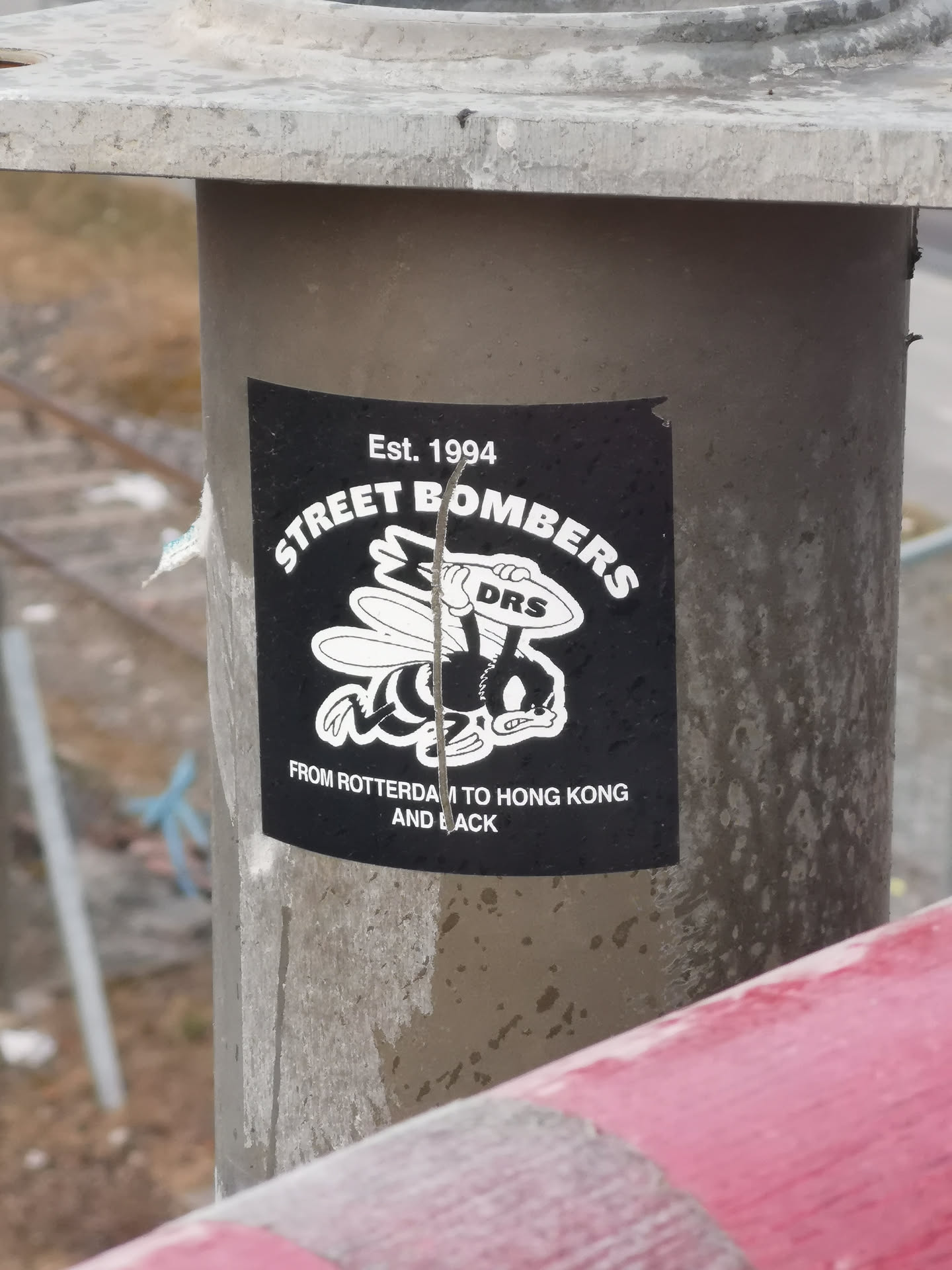 The sticker is a black rectangle with slightly rounded corners, featuring white text and a cartoon graphic. The central image is a stylized, angry-looking cartoon bee, wearing gloves and holding an oval object with 'DRS' written on it. Above the bee, in an arched font, is 'STREET BOMBERS'. At the very top, centered, it reads 'Est. 1994'. Below the bee, in two lines, it states 'FROM ROTTERDAM TO HONG KONG' and 'AND BACK'. The sticker appears to be affixed to a metal pole and shows some signs of wear, including a vertical tear or scratch down its center.