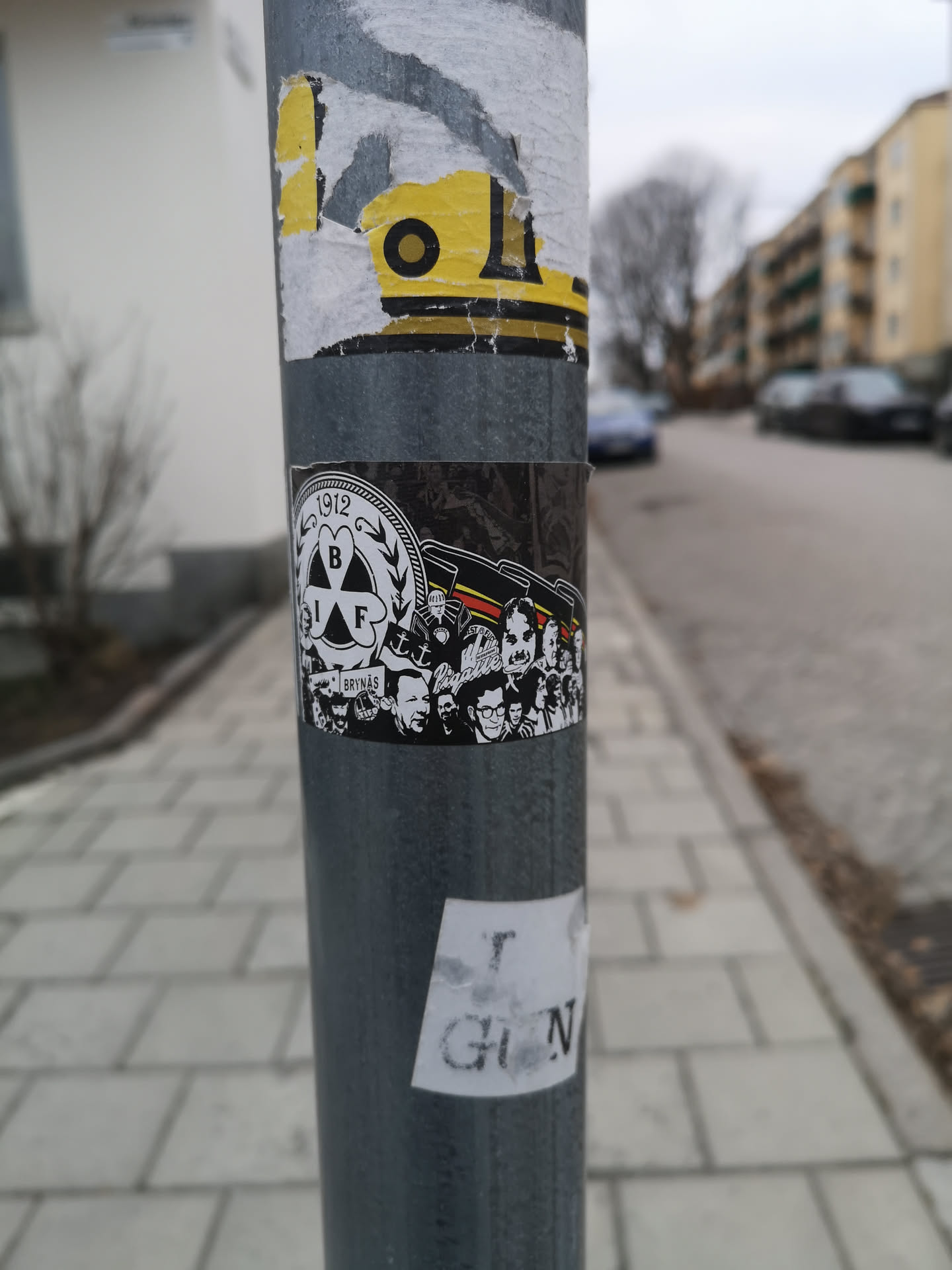A rectangular sticker adhered to a metal pole, featuring a black and white design with accents of red and yellow. The left side displays a circular crest with '1912' at the top, 'BIF' in the center within a shamrock-like emblem, and 'BRYNÄS' below, all in white on a black background. The right side depicts a stylized crowd of diverse faces, rendered in black and white, with several red and yellow flags or banners waving above them. The overall aesthetic is graphic and illustrative, celebrating a sports club's history and supporters.