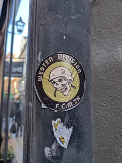 A round sticker slapped onto a dark vertical metal pole. It features a central yellow/gold circle depicting a stylized human skull wearing an army helmet with an unbuckled chinstrap. Encircling this graphic is a thick black band with stylized gothic lettering that reads 'WESTEN DIVISION' across the top and 'F.C.M. ZH' at the bottom. The surface of the sticker is slightly wrinkled and weathered.