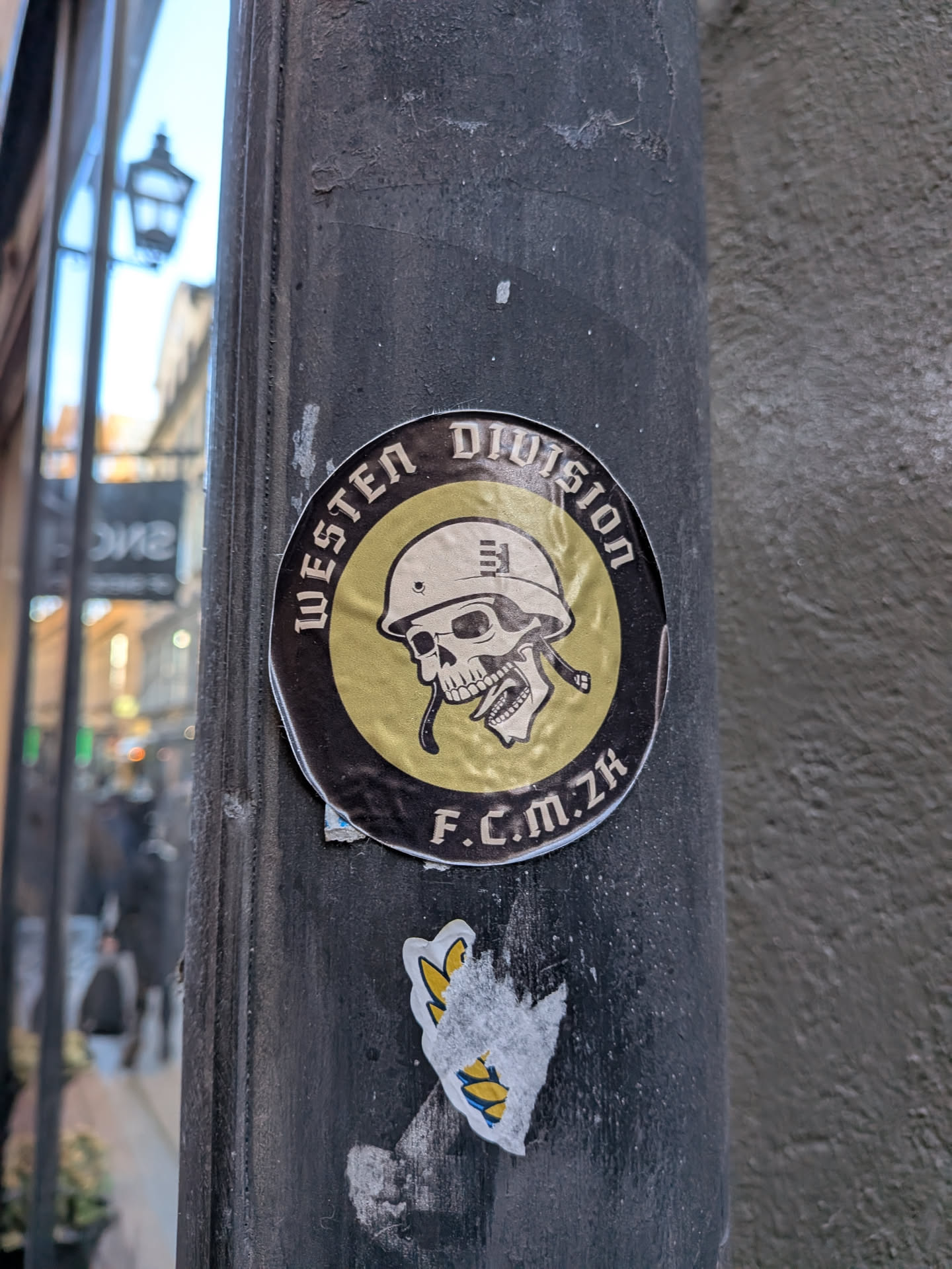 A round sticker slapped onto a dark vertical metal pole. It features a central yellow/gold circle depicting a stylized human skull wearing an army helmet with an unbuckled chinstrap. Encircling this graphic is a thick black band with stylized gothic lettering that reads 'WESTEN DIVISION' across the top and 'F.C.M. ZH' at the bottom. The surface of the sticker is slightly wrinkled and weathered.