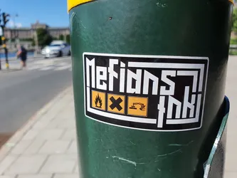 A black and white sticker with the text "Metians Ink" in a stylized font. It also features three small hazard symbols: fire, biohazard, and corrosive. The sticker is affixed to a green pole.